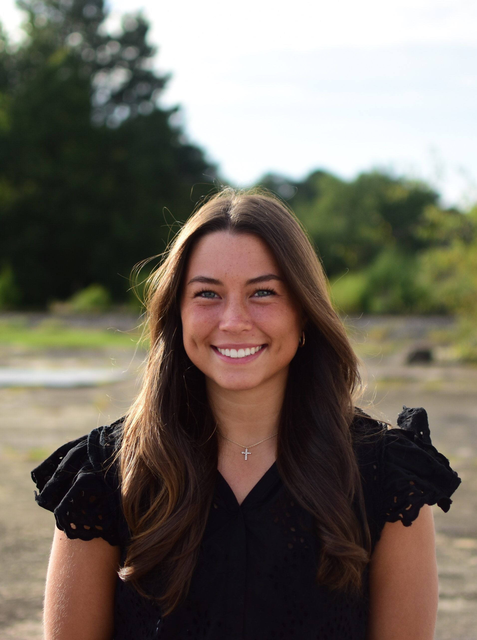 A person with long brown hair, wearing a black top and a small cross necklace, smiles in front of a blurred green field.
