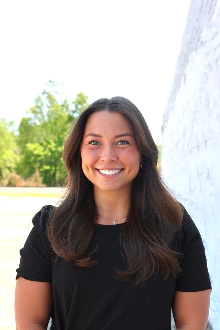 A person with long brown hair, wearing a black top and a small cross necklace, smiles in front of a blurred green field.