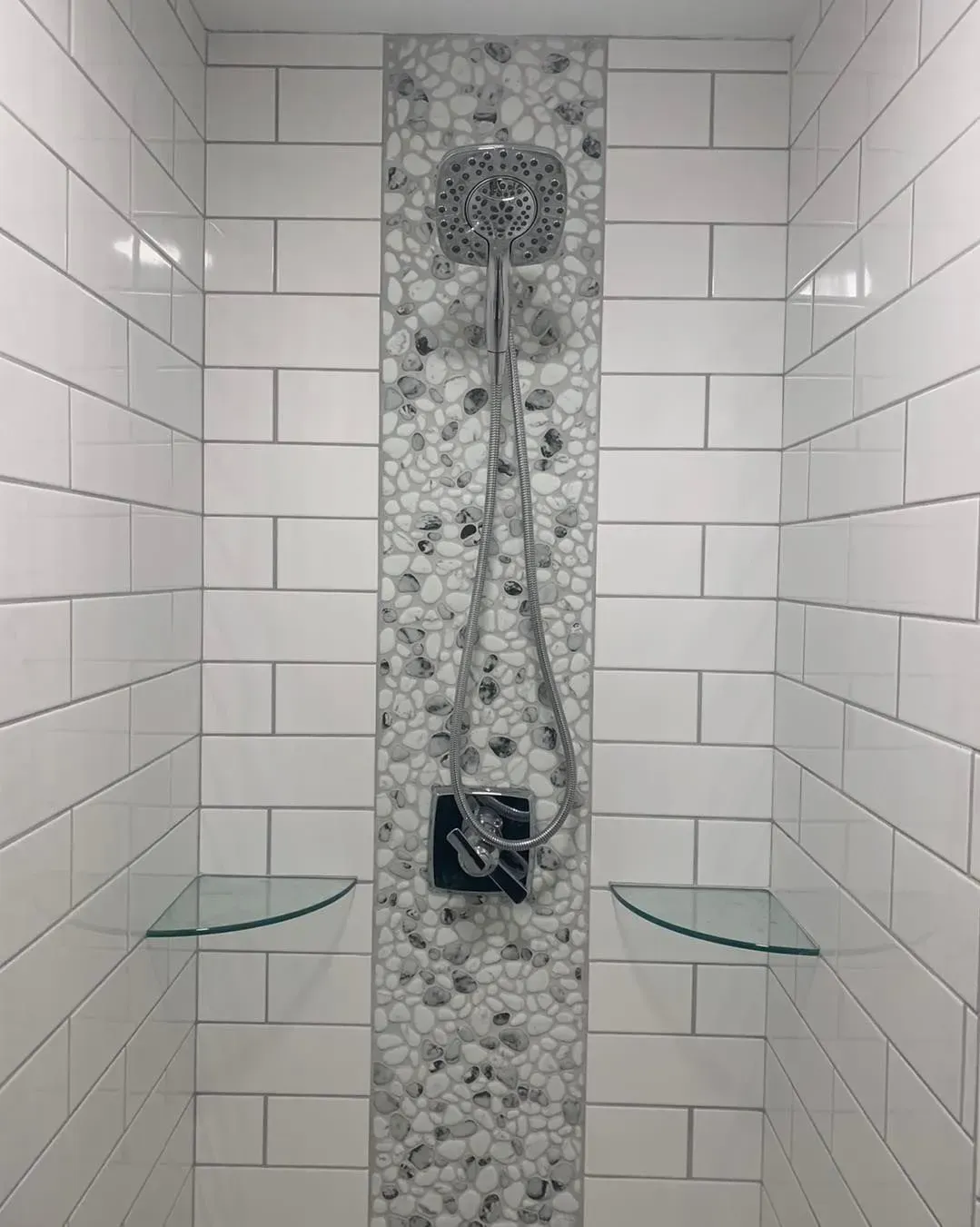 A shower with white tiles and glass shelves and a shower head.