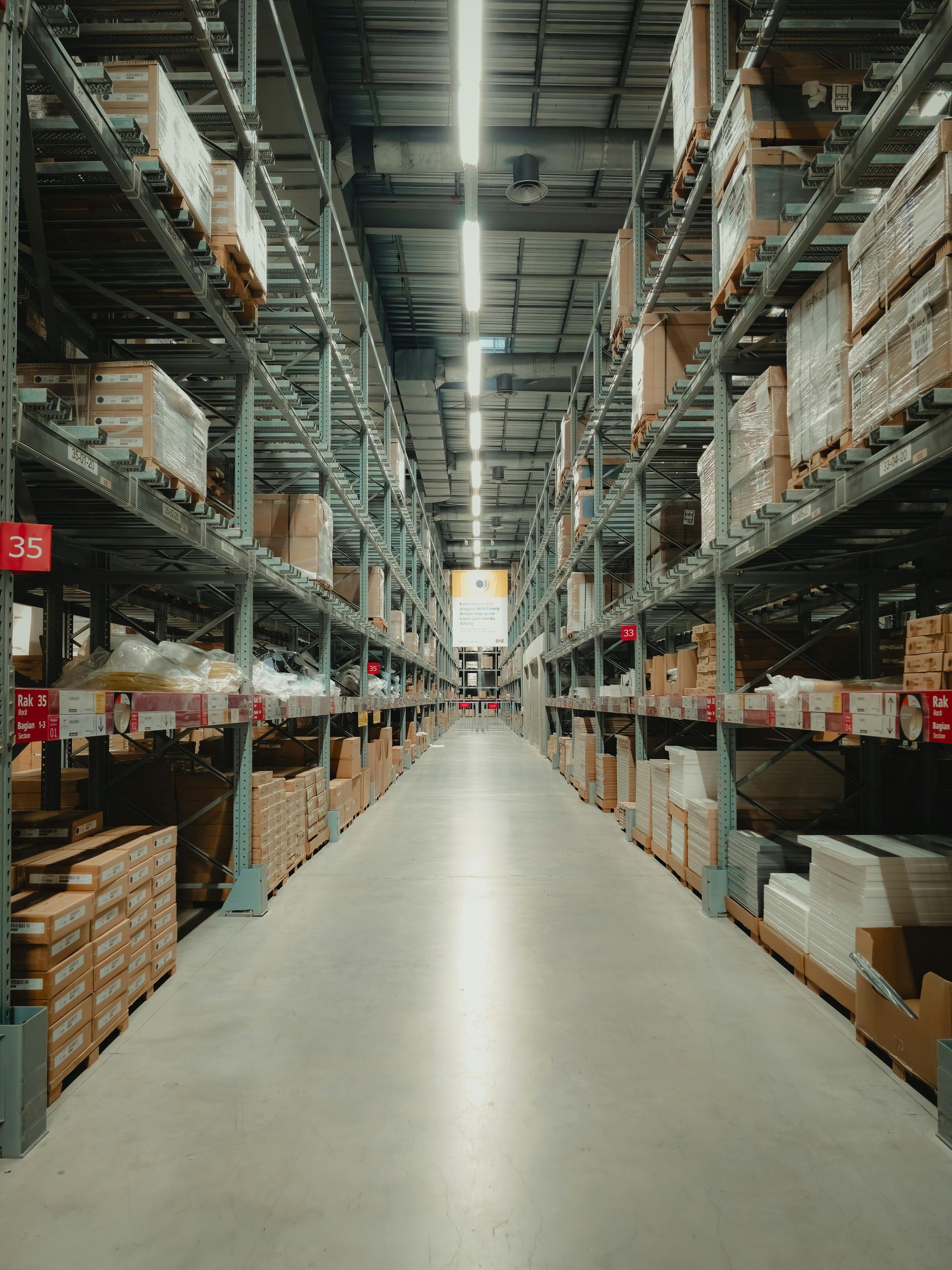 A long, brightly lit warehouse aisle with tall metal shelves stacked with cardboard boxes on both sides.  Epoxy floor