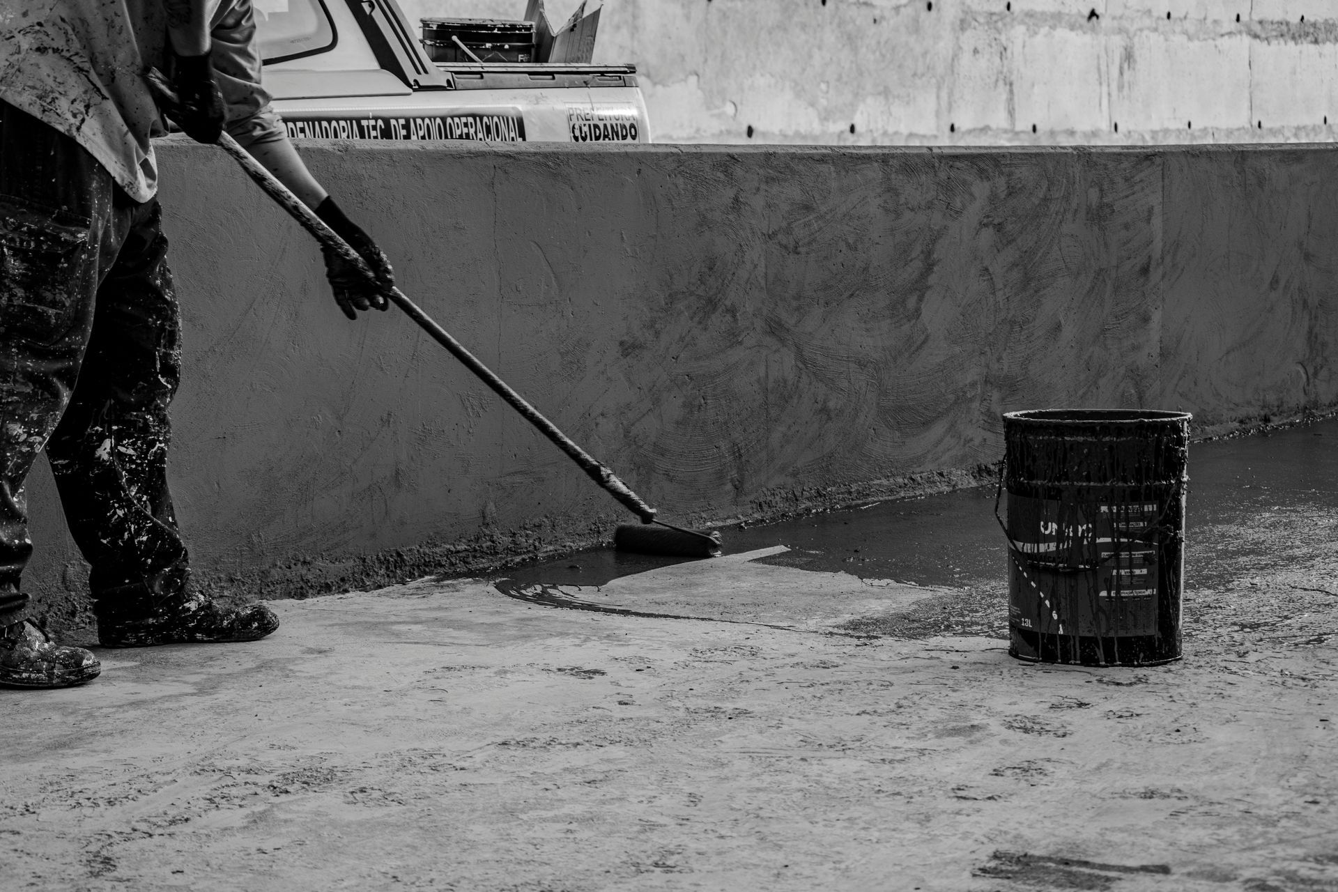 A person uses a long-handled brush to apply dark liquid coating to a concrete floor near a bucket and wall.  Epoxy floor