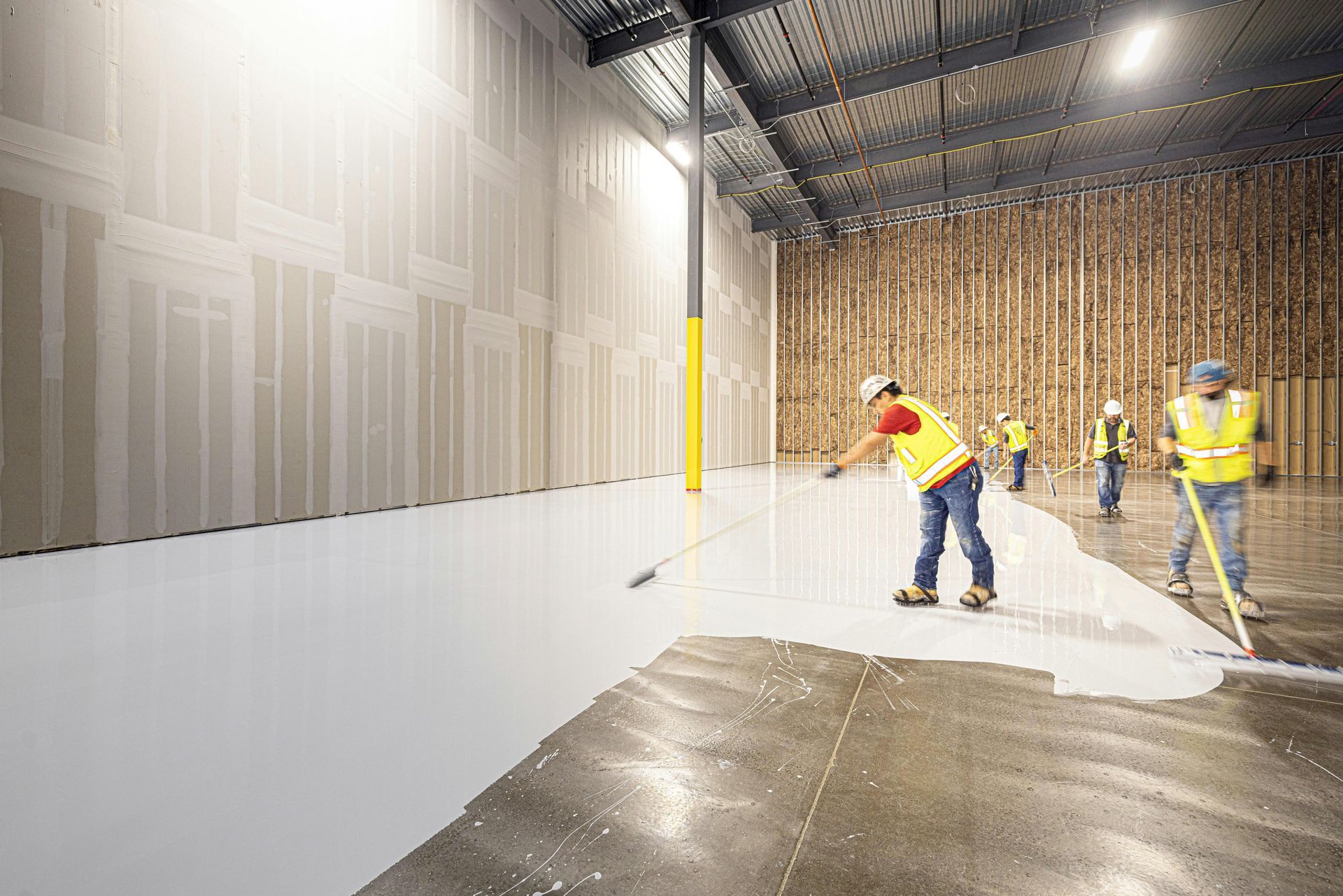 Workers in safety vests apply a white epoxy coating to a large concrete warehouse floor.