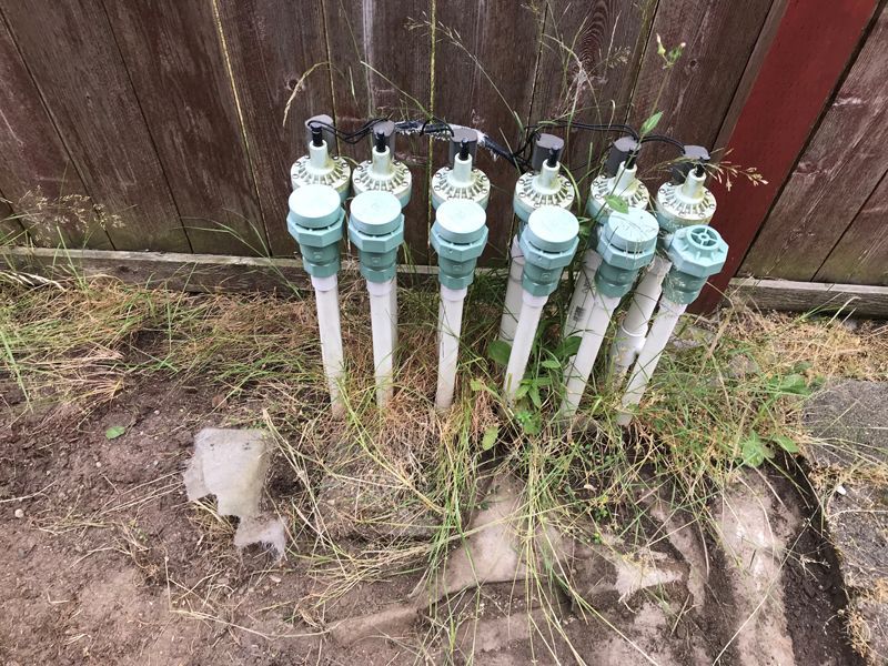 Green sprinkler heads with beige and white pipes emerge from the ground near a wooden fence.