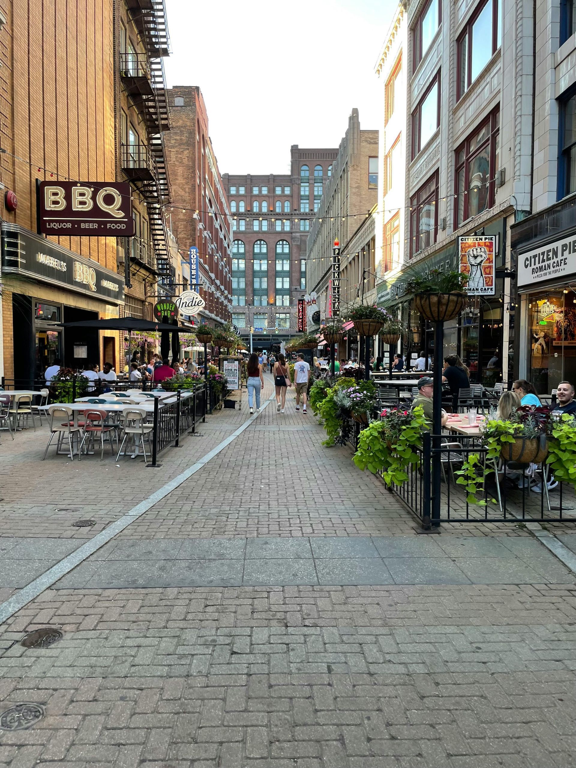 Brick-paved pedestrian street lined with restaurants, outdoor seating, and tall buildings in an urban setting.