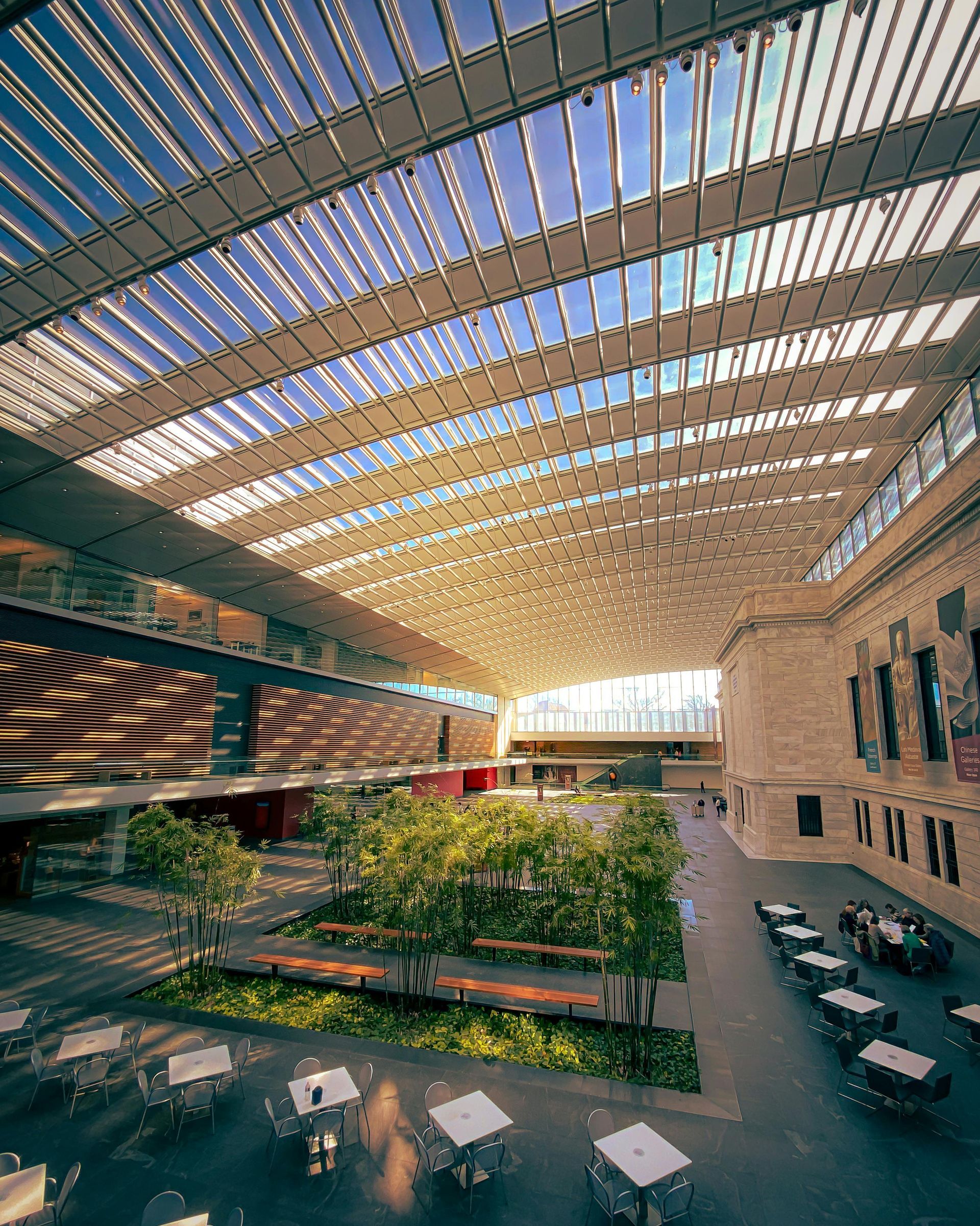 A wide, open interior courtyard with a glass roof. Trees, seating, and outdoor cafe tables are in the foreground.