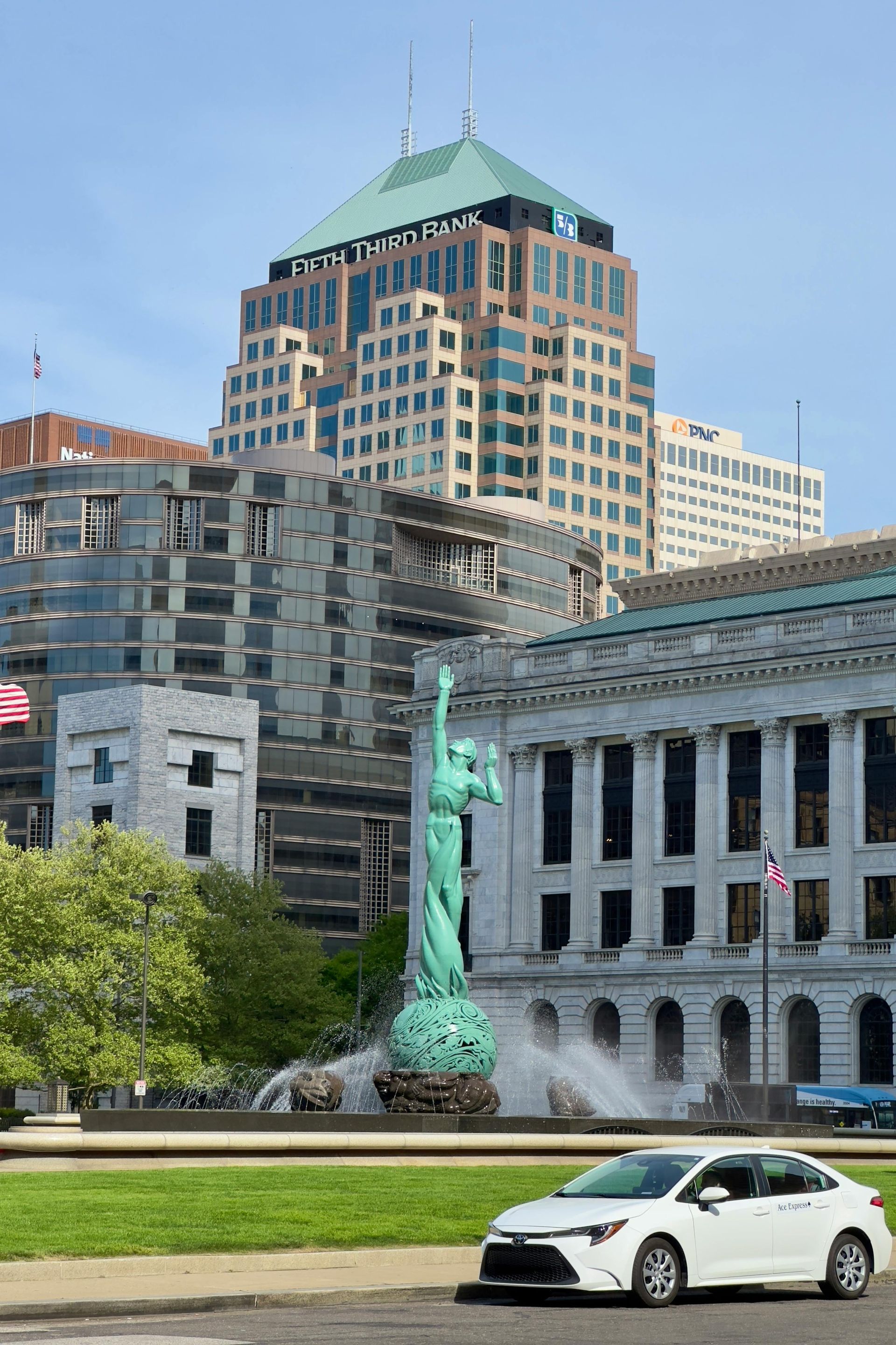 White sedan in front of a green statue and building with a modern high-rise in the background, Cleveland, Ohio.