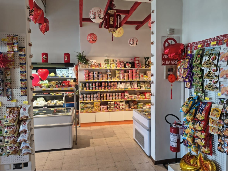 Interno di un negozio di alimentari asiatico con scaffali pieni di prodotti confezionati, banchi frigo e decorazioni festive con lanterne di carta rosse.