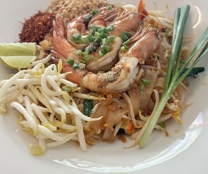Un piatto di Pad Thai con gamberi, guarnito con germogli di soia, frutta secca tritata, peperoncino in polvere, uno spicchio di lime e cipollotti.