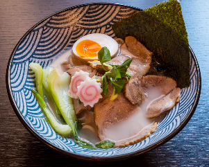 Una ciotola di ramen con fettine di maiale, un uovo alla coque, tortini di pesce, bok choy e alga nori in una ciotola con motivo blu.