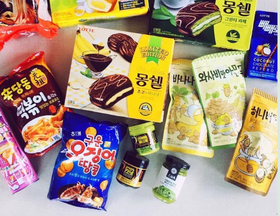 Una collezione di snack coreani, tra cui tortine Lotte Mongshell, gnocchi di riso piccanti e varie patatine in busta, disposti su una superficie bianca.