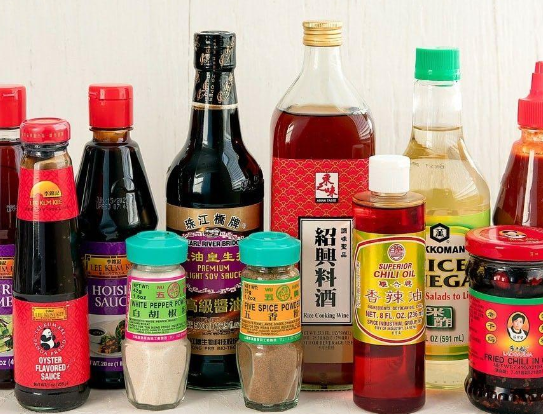 Una collezione di diverse salse, aceti e spezie asiatiche in bottiglie e barattoli, disposte su uno sfondo bianco.