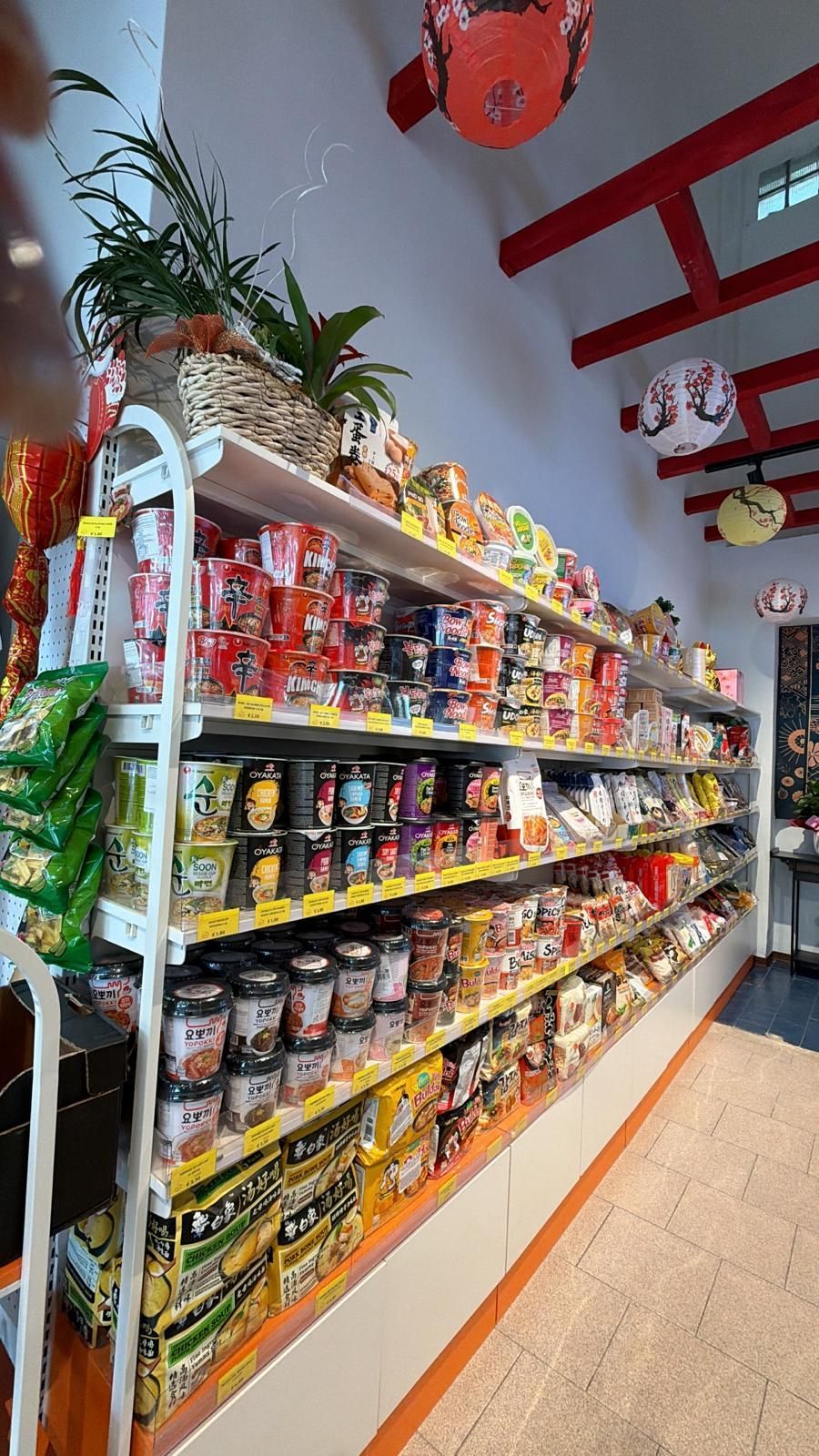 Un corridoio di un negozio con scaffali pieni di vari tipi di noodles istantanei in tazza e in confezione, illuminati da lanterne decorative appese.