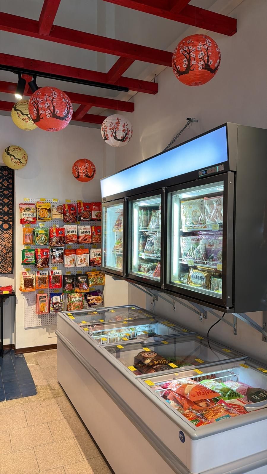 Una corsia di un supermercato con un congelatore a pozzetto e frigoriferi verticali con porte in vetro, decorata con lanterne di carta sospese.