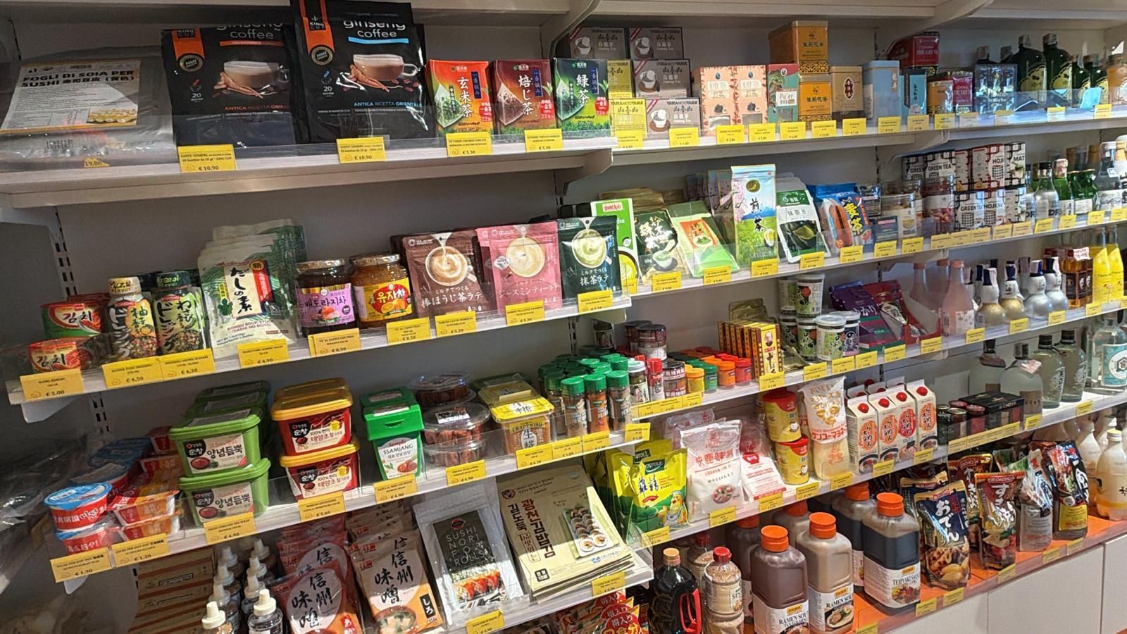 Scaffali di un mercato pieni di vari prodotti alimentari confezionati, tra cui snack, salse e vasetti, con etichette di prezzo gialle.