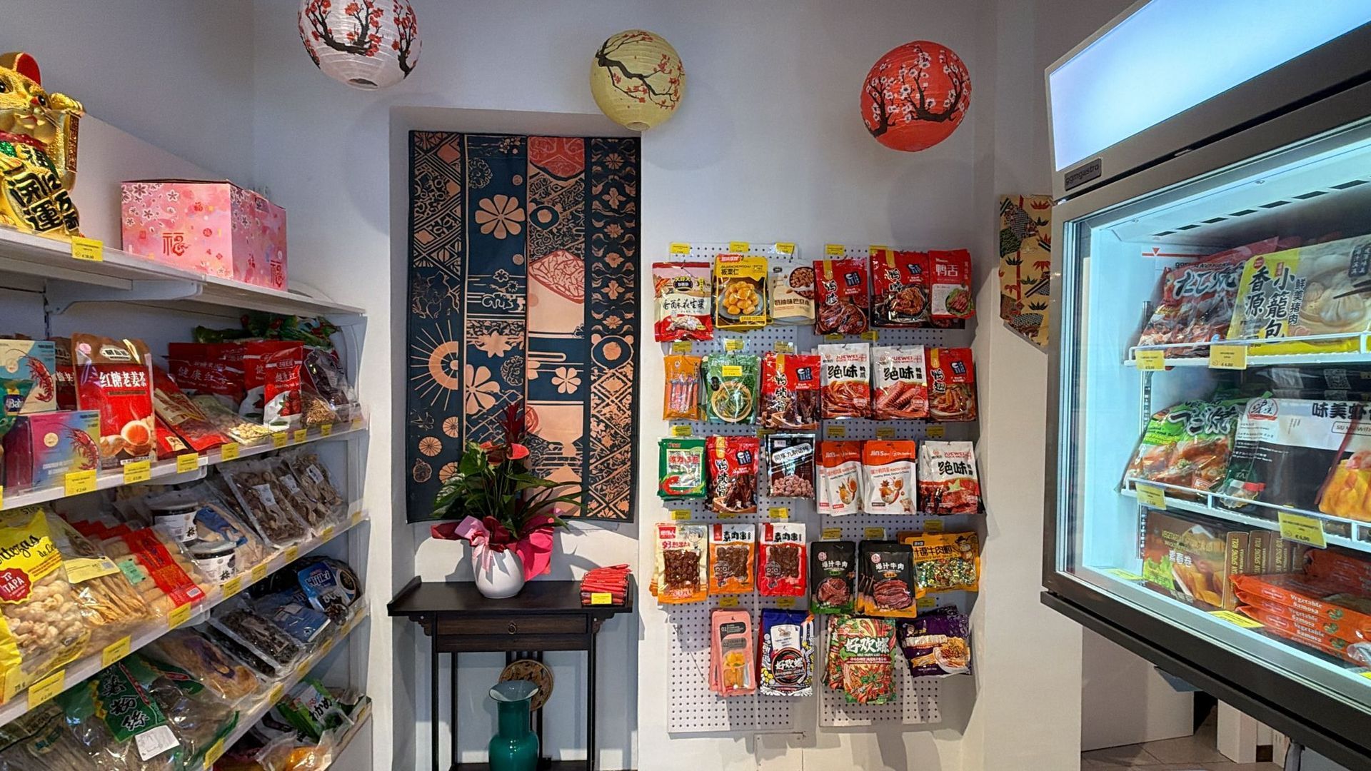 L'interno di un piccolo negozio con scaffali per snack, espositori per snack appesi, un arazzo decorativo alla parete e un frigorifero.