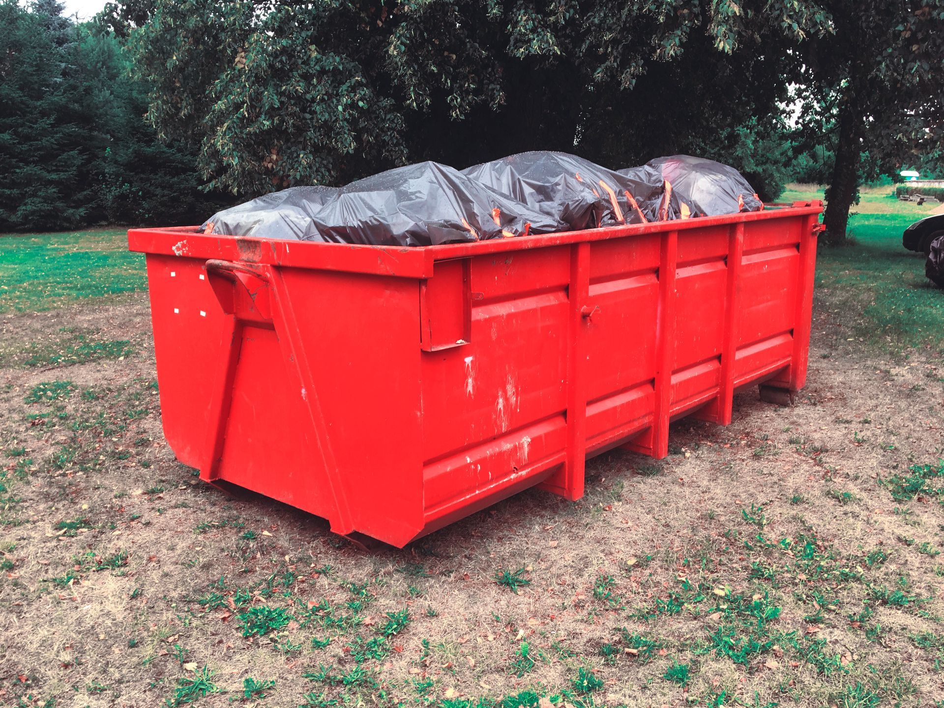 Bright red dumpster filled with covered trash bags on a grassy outdoor lot