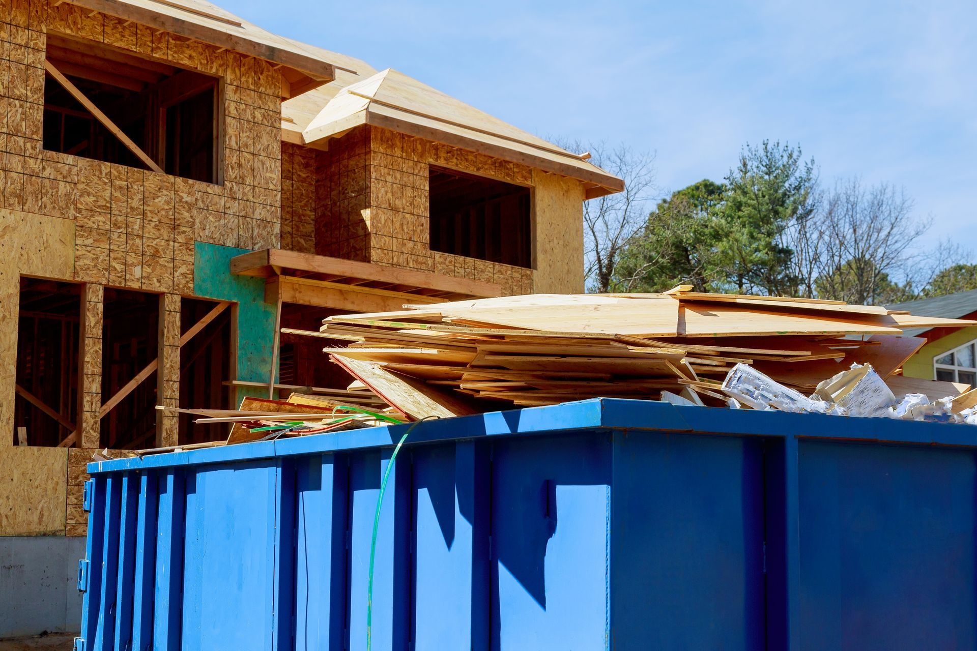 Partially built wooden house behind a blue dumpster with construction debris on top
