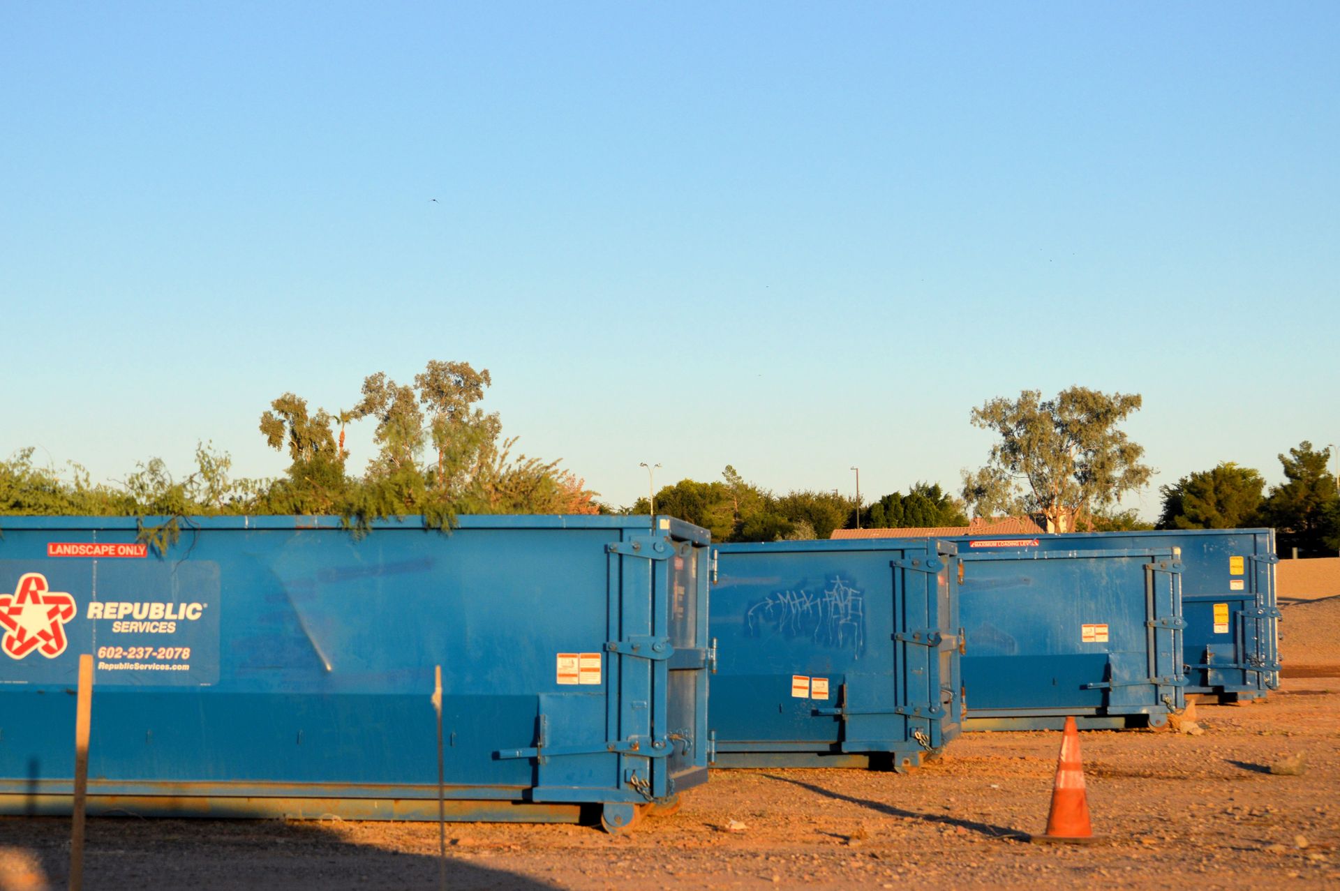 Blue construction dumpsters lined up on a dirt lot at sunset, with trees and an orange cone nearby