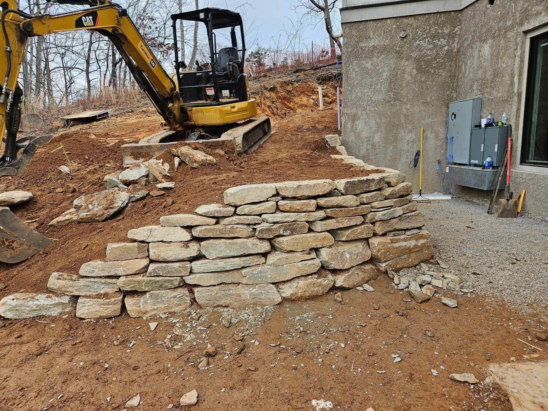 A yellow excavator is working on a stone wall in front of a house.