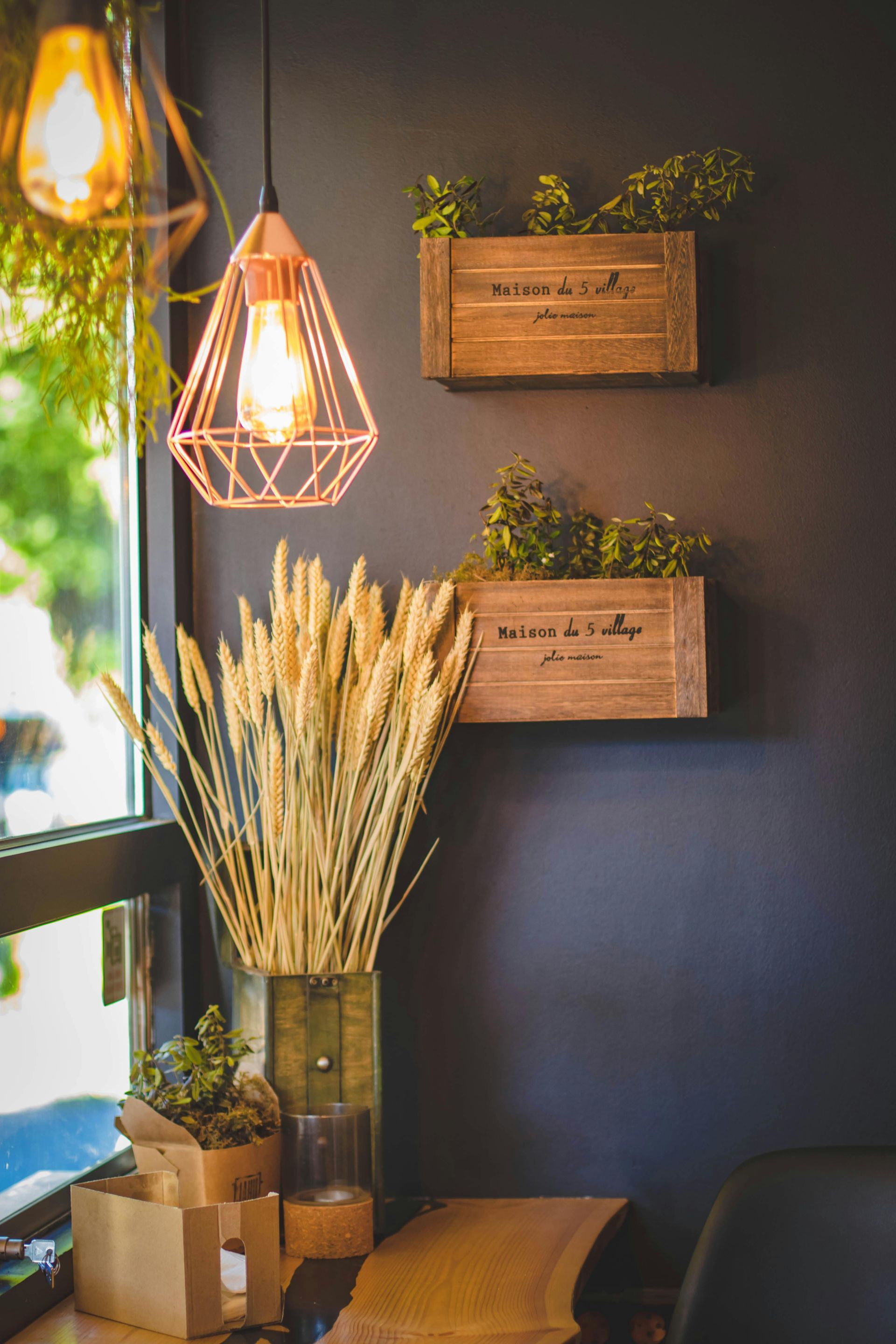 Interior cafe scene: gold light, wooden boxes with greenery, wheat stalks, dark wall, window.