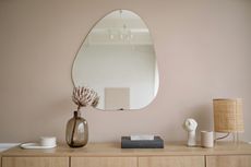 Beige wall with an irregular mirror, floral arrangement, and decorative items on a wooden sideboard.