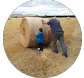 An adult and a small child push a large, round hay bale across a dry, grassy field.