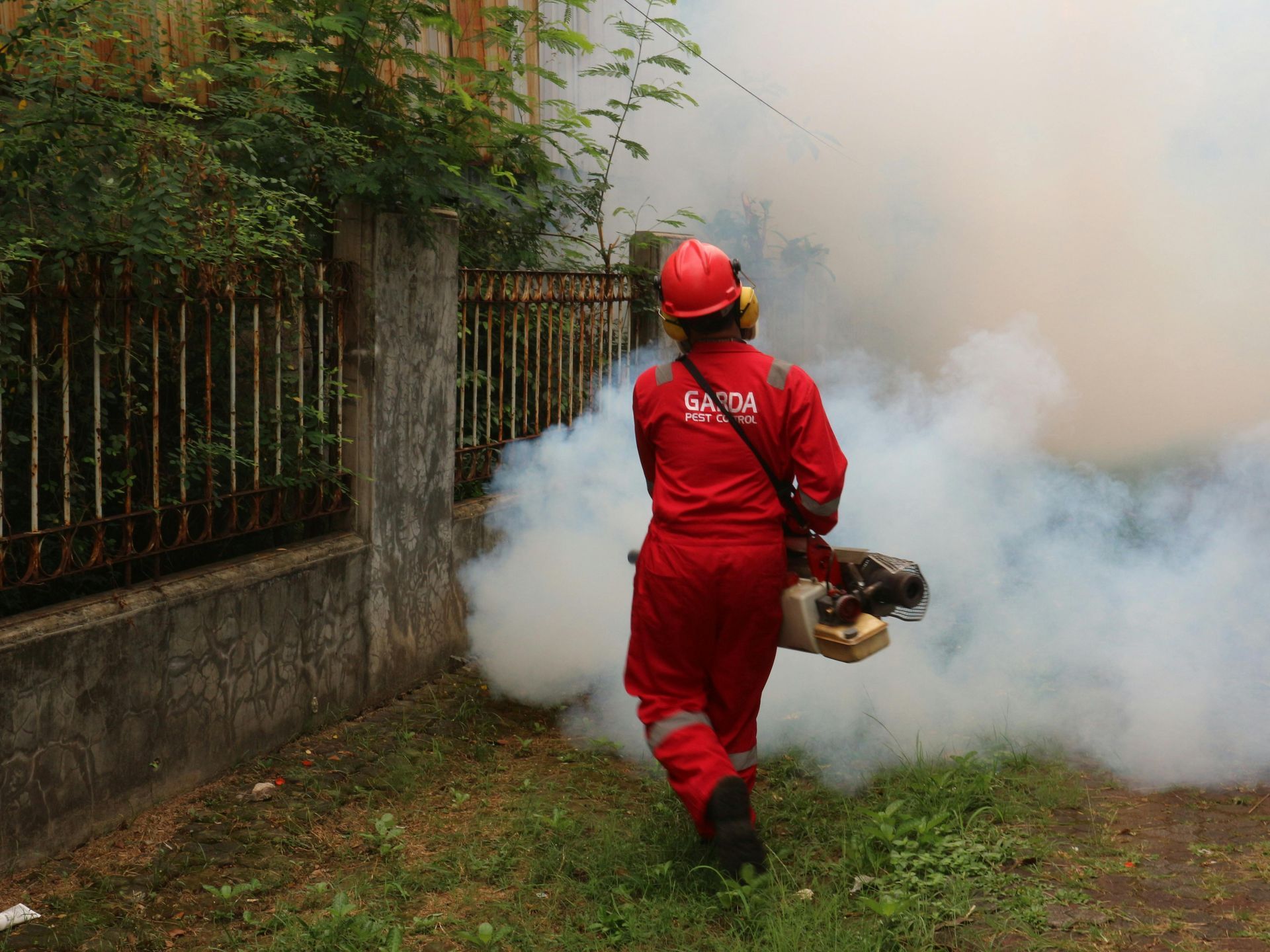 A worker in a red uniform walks alongside a fence while operating a portable fogging machine to spray insecticide.