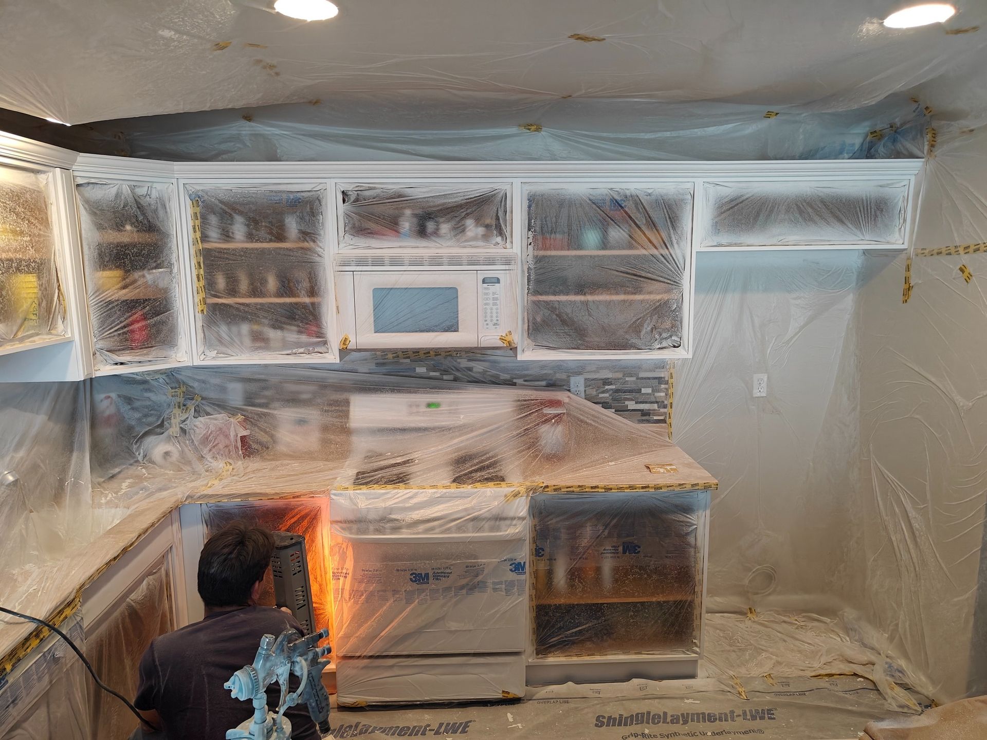 A man is painting a kitchen with plastic covering the cabinets.