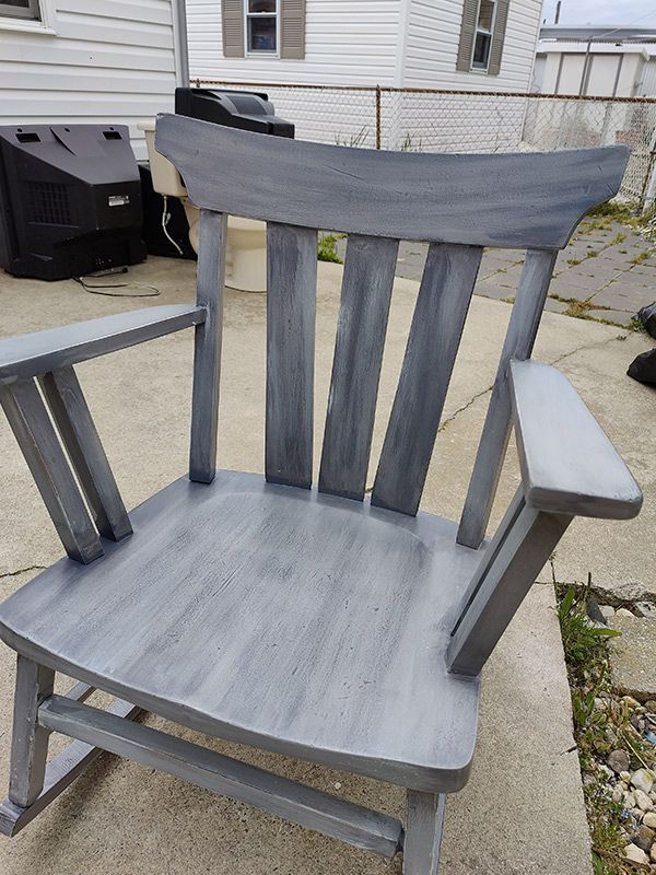 A gray wooden rocking chair is sitting on a sidewalk in front of a house.