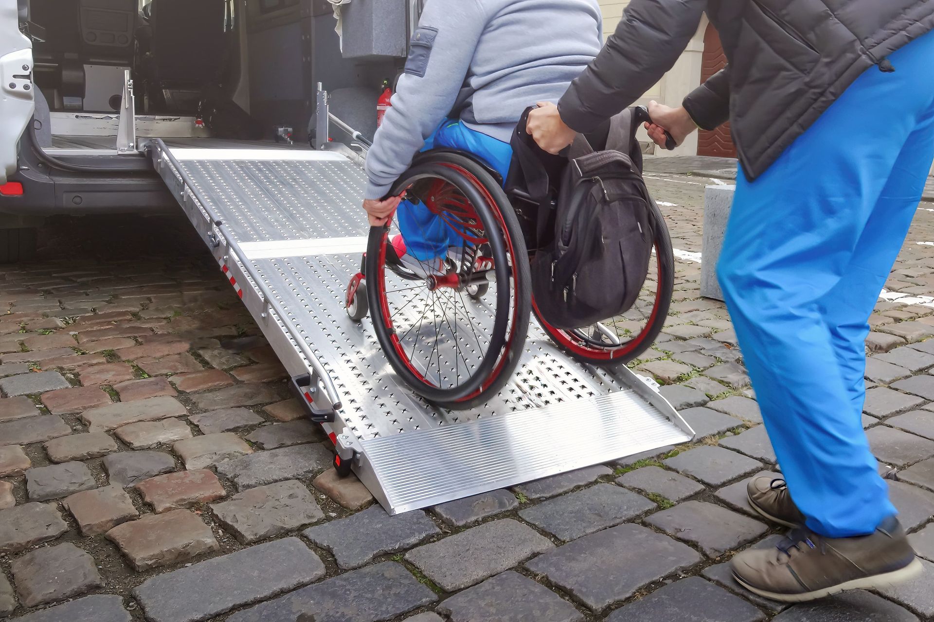 Person in a wheelchair being pushed up a metal ramp from a van onto a cobblestone street
