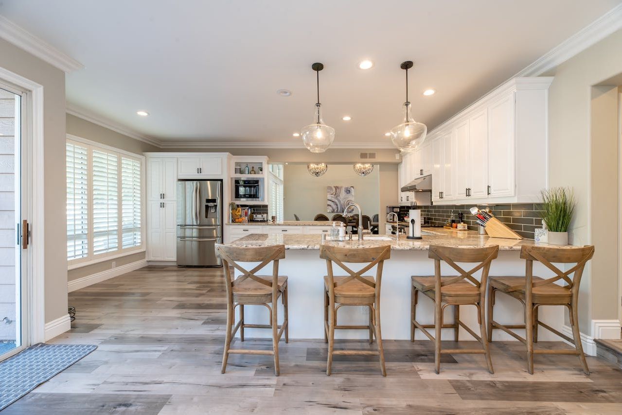 A kitchen with white cabinets , wooden floors , stainless steel appliances and a large island with chairs.
