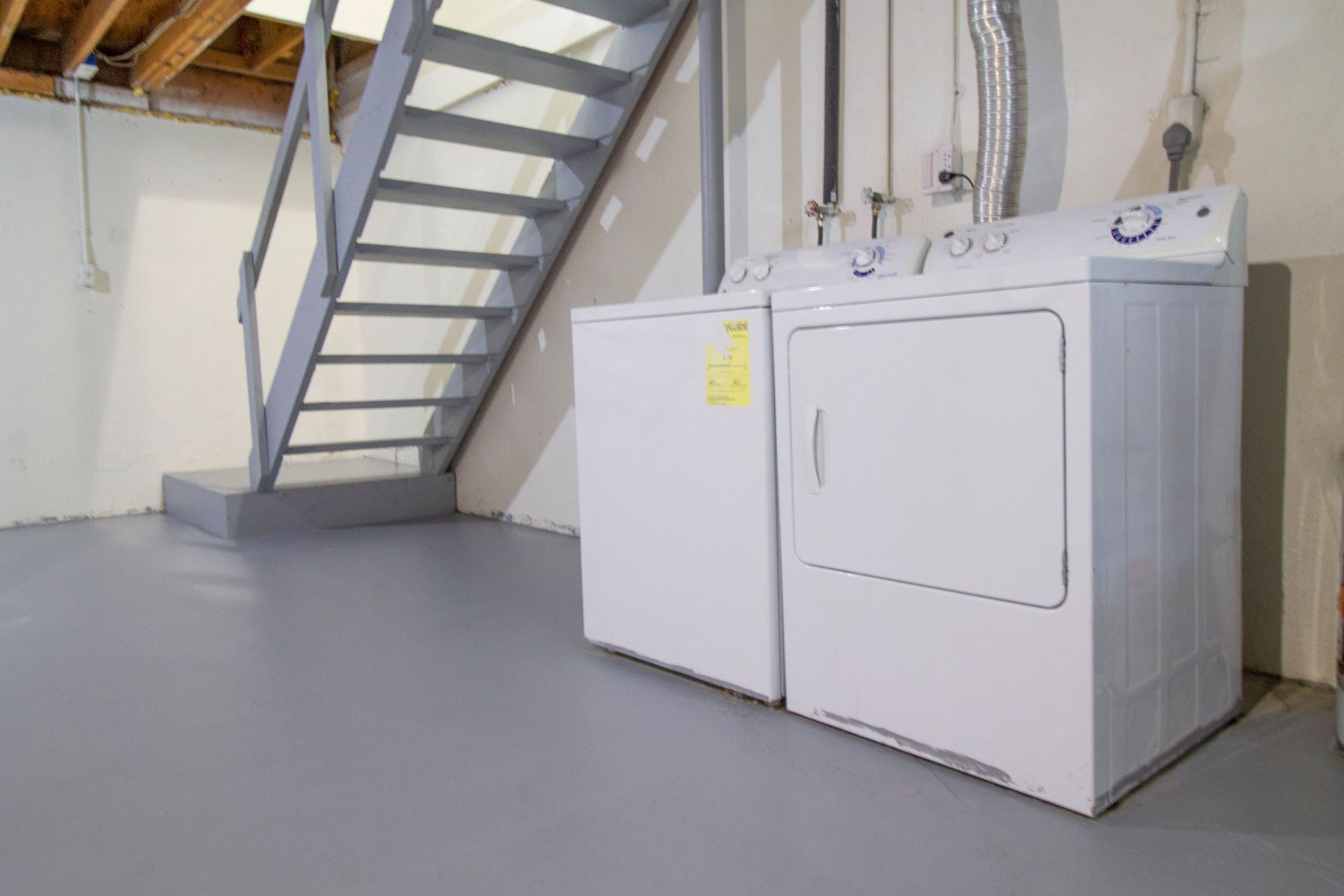 A washer and dryer are sitting in a basement next to a staircase.