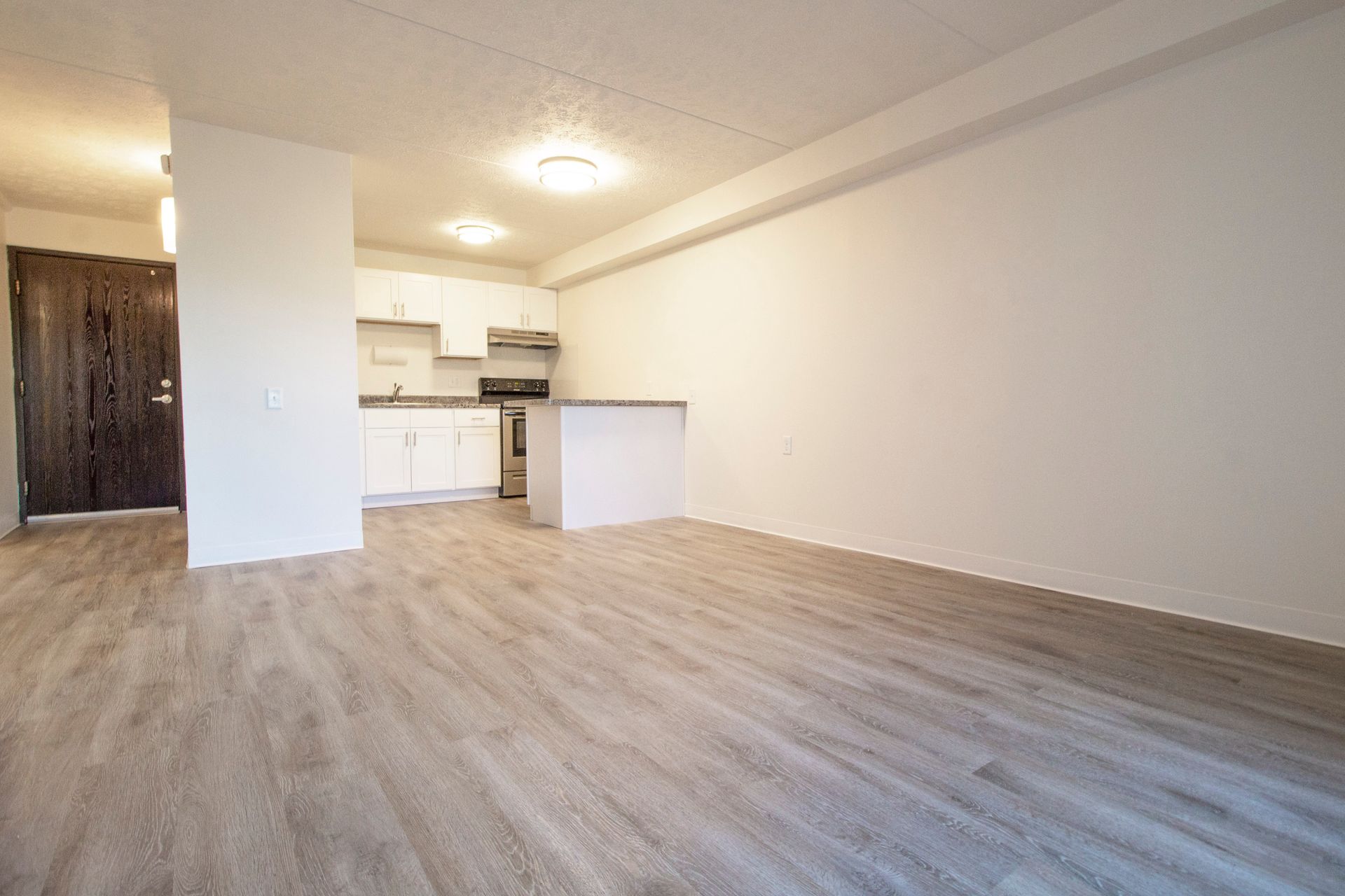 An empty living room with hardwood floors and a kitchen in the background.