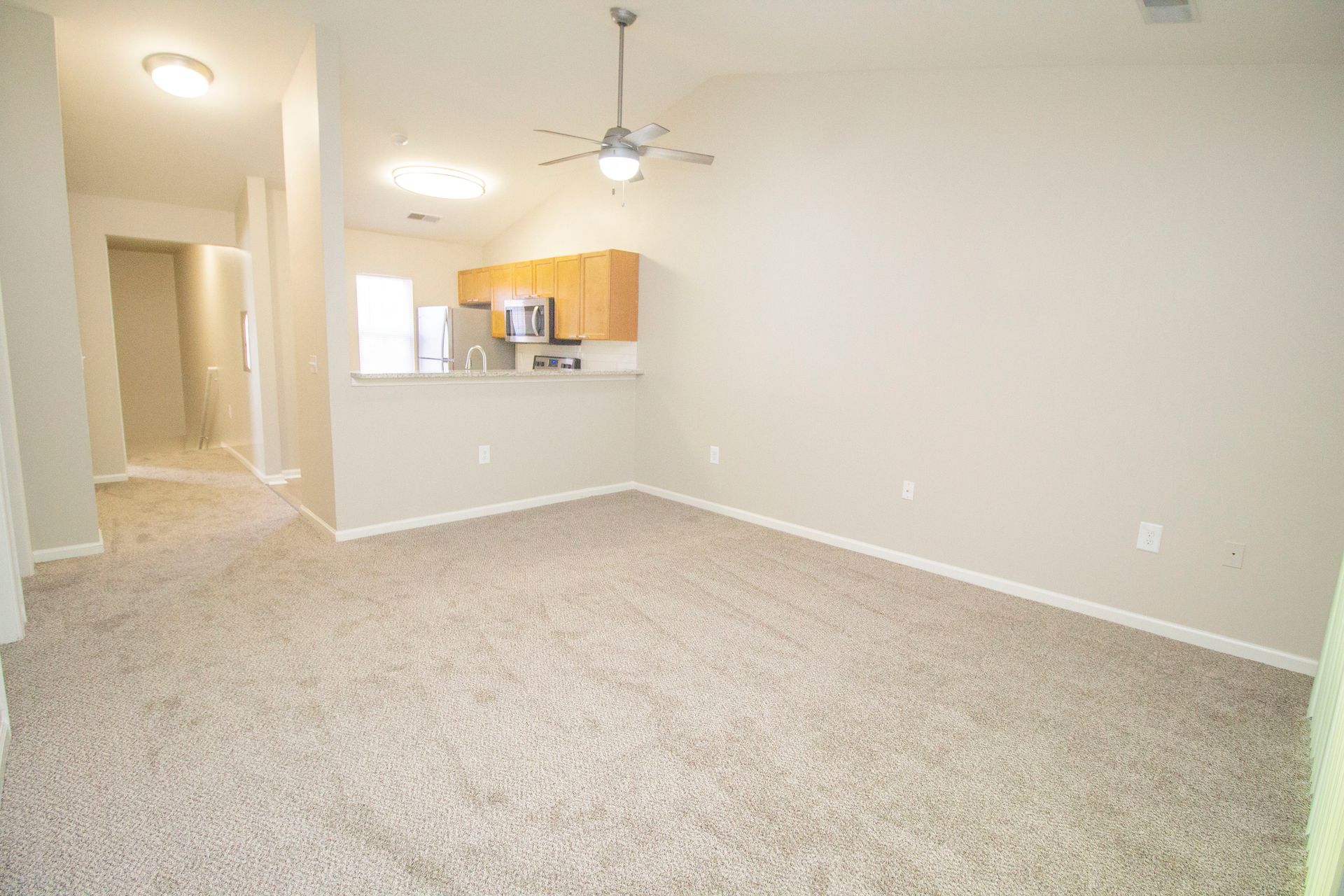 An empty living room with a ceiling fan and a kitchen in the background.