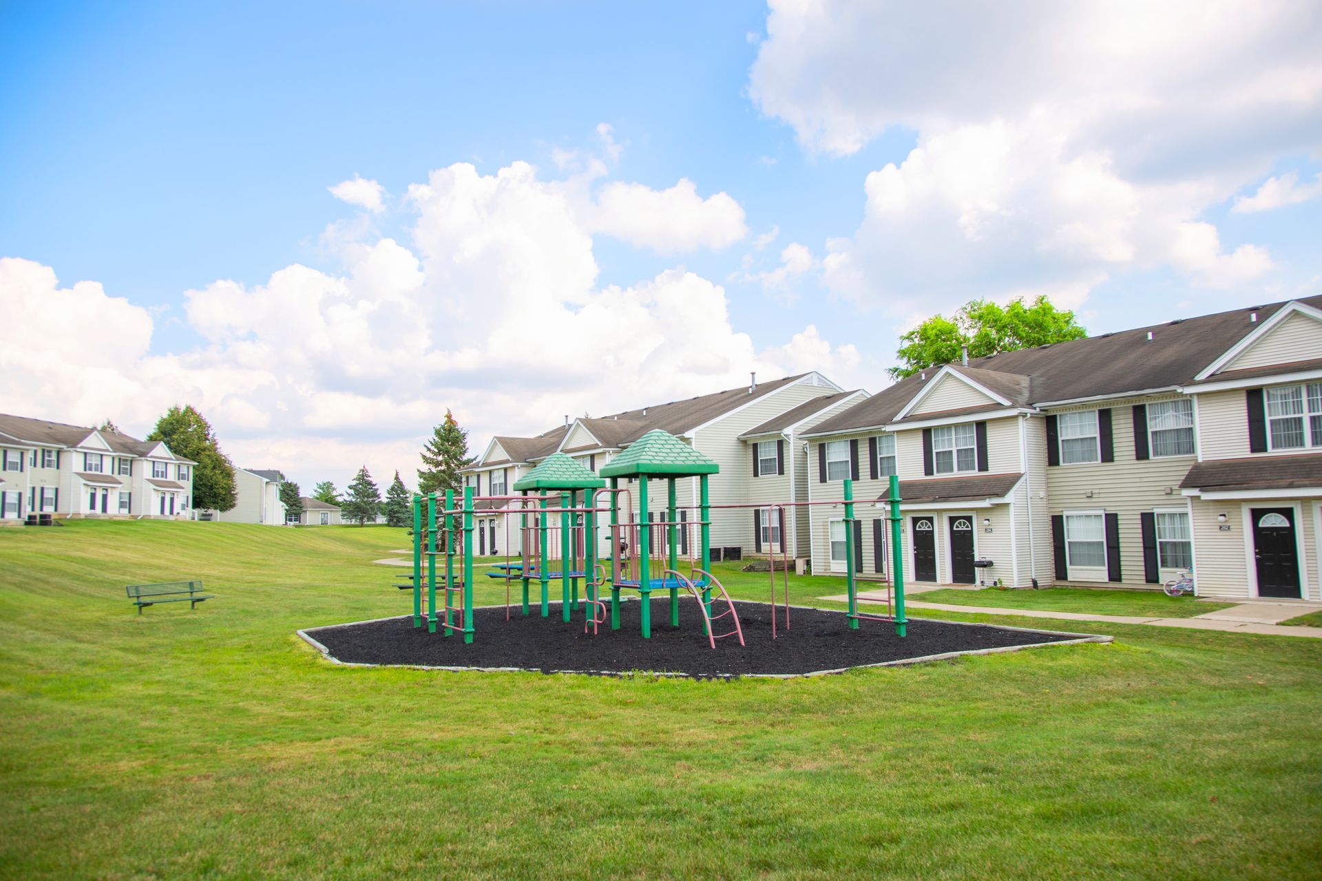 A playground is in the middle of a lush green field in front of a row of houses.