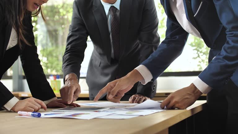 Business people in suits gather, pointing at papers on a table.
