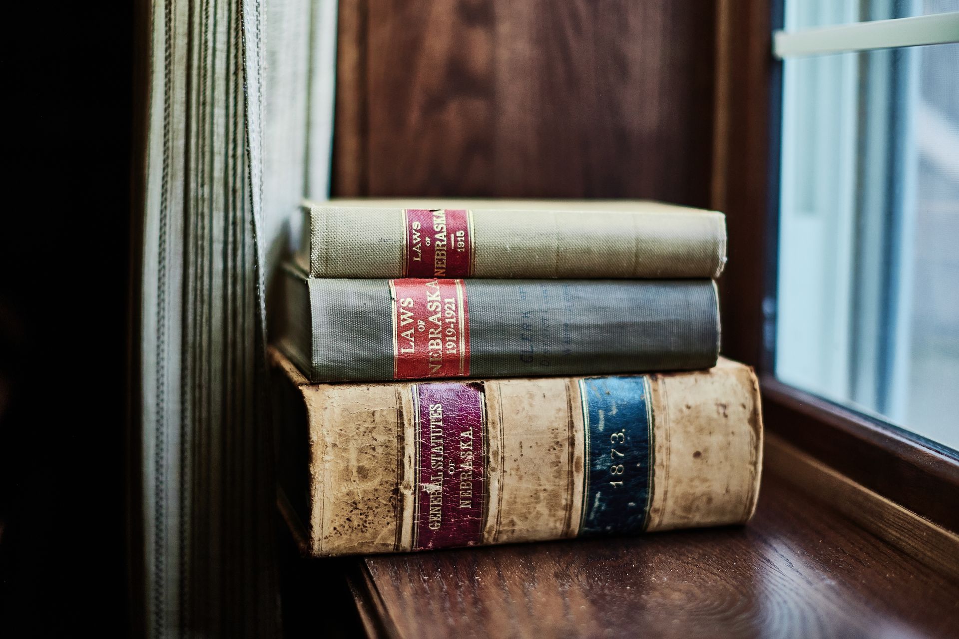 Three stacked antique books on a dark wooden windowsill, with colorful spine labels.