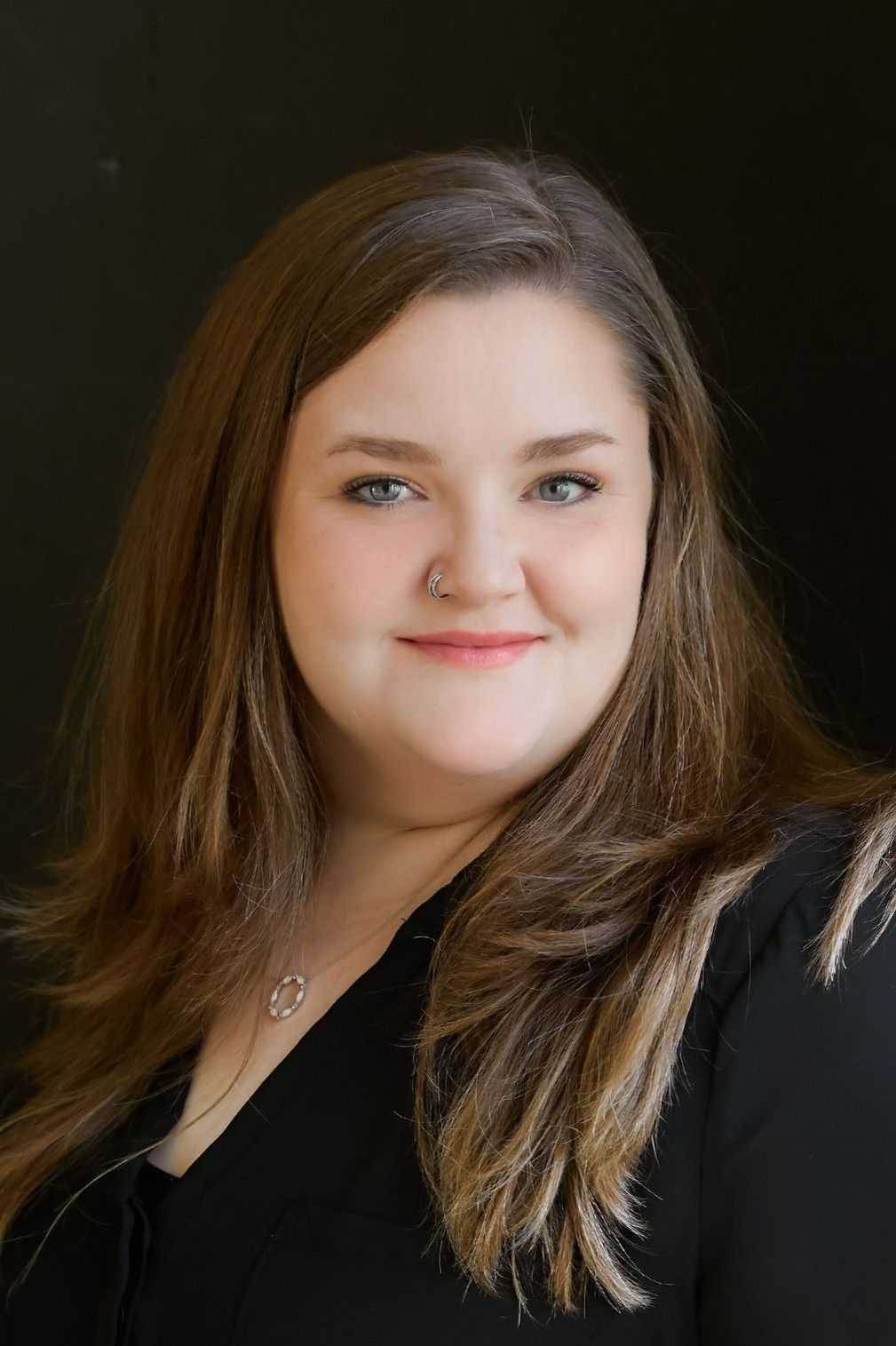 Woman smiles at the camera, wearing a blue top and black jacket, indoors, wood background.