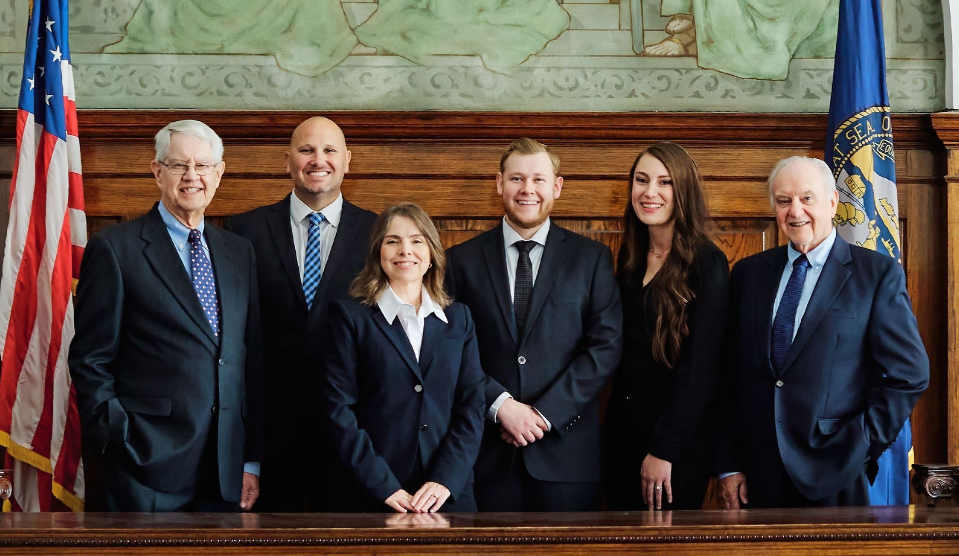 Six people in suits stand behind a wooden desk, in front of wood paneling and a painted mural.