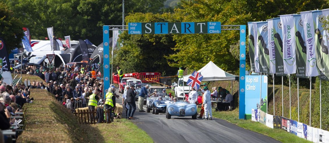 Cars lined up at the starting line of a race, under an archway reading 
