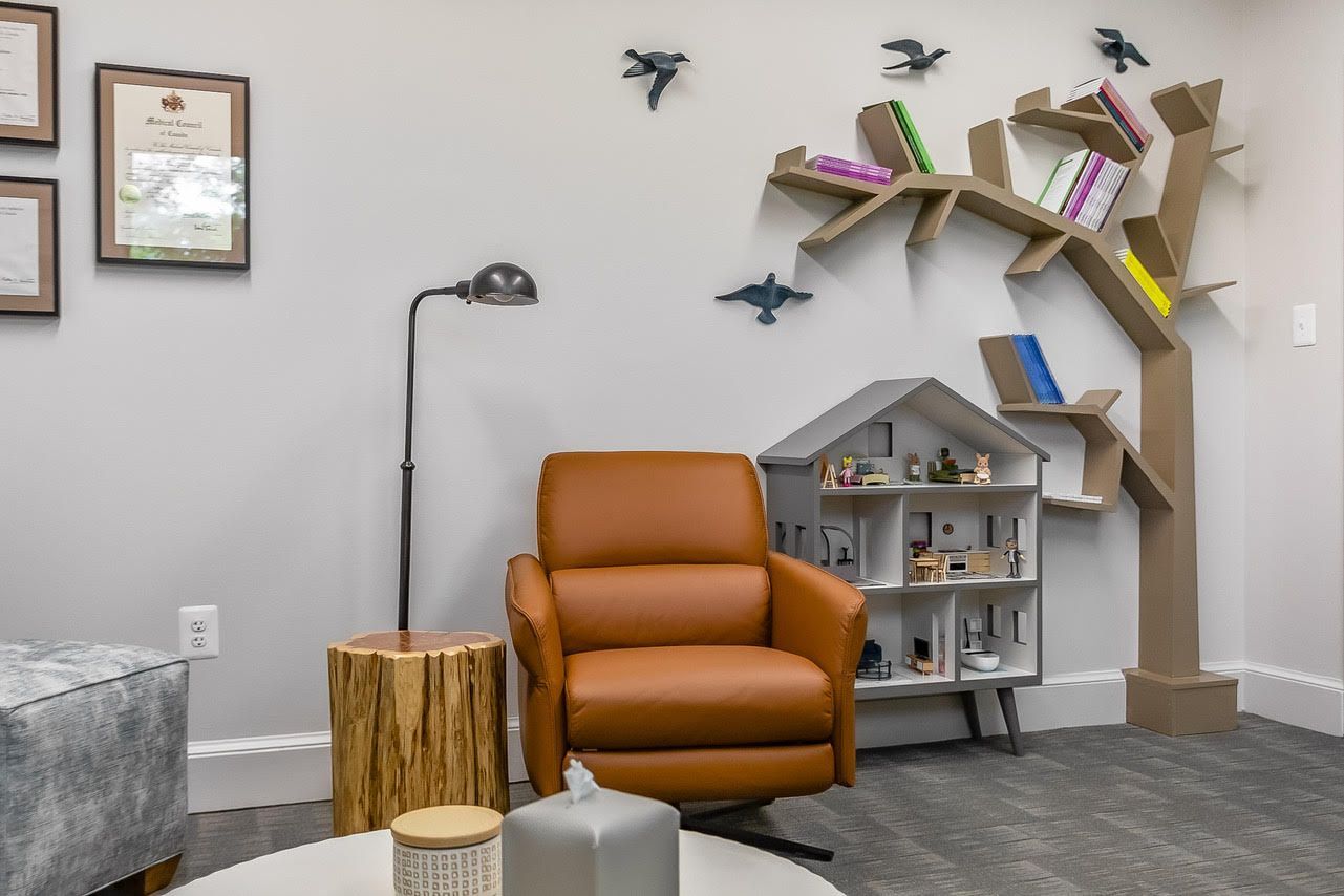 Dr Sehon's therapy  room with a brown chair , a table , a lamp, a tree shaped bookshelf and a doll house.