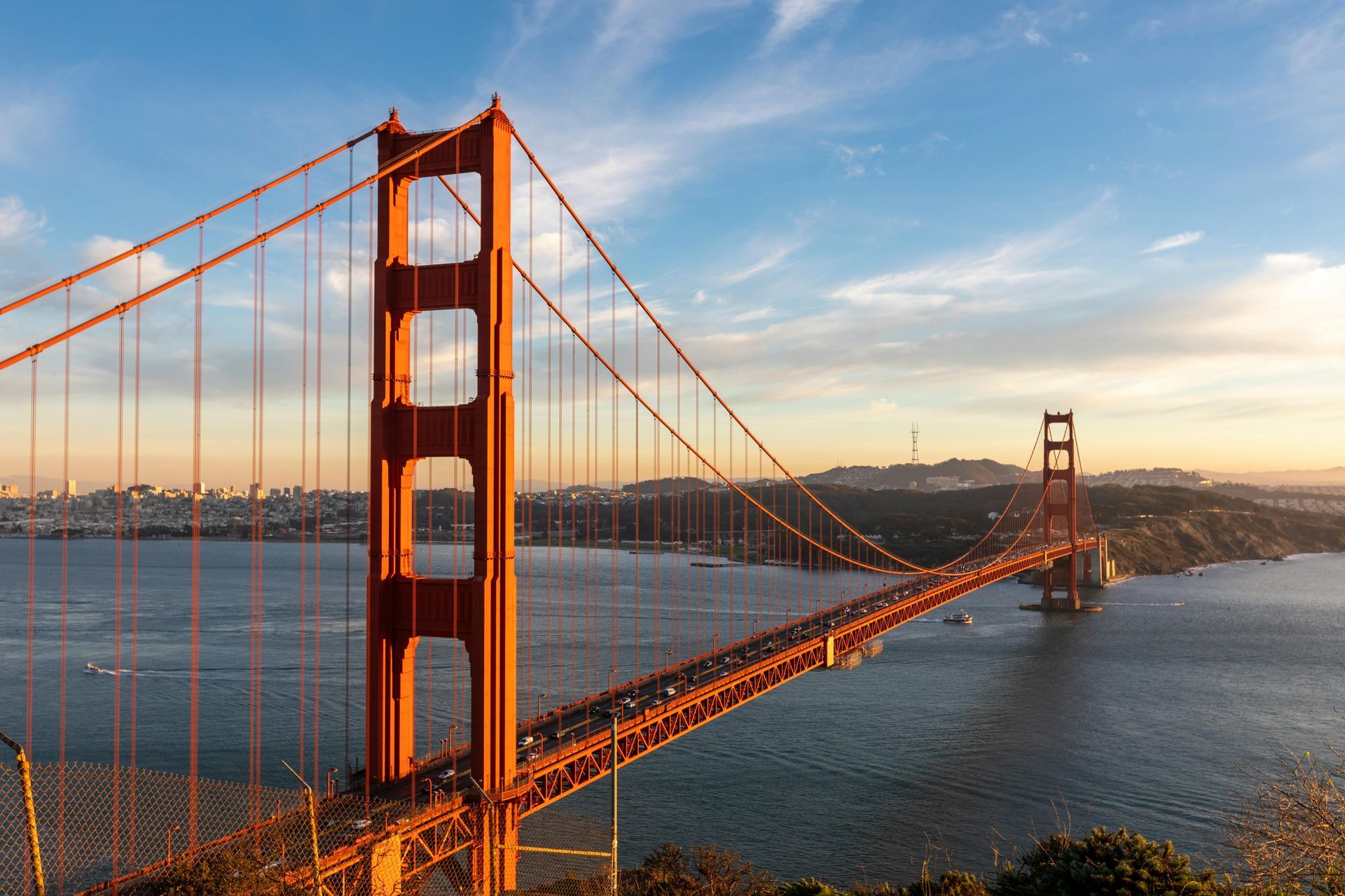 Golden Gate Bridge at sunset, with its iconic orange towers and cables stretching across the water. City skyline in background.