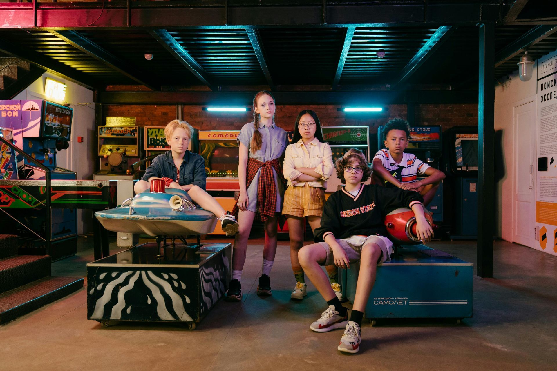 Group of five young people posing in an arcade. They are surrounded by arcade games, wearing casual clothing, and looking towards the camera.