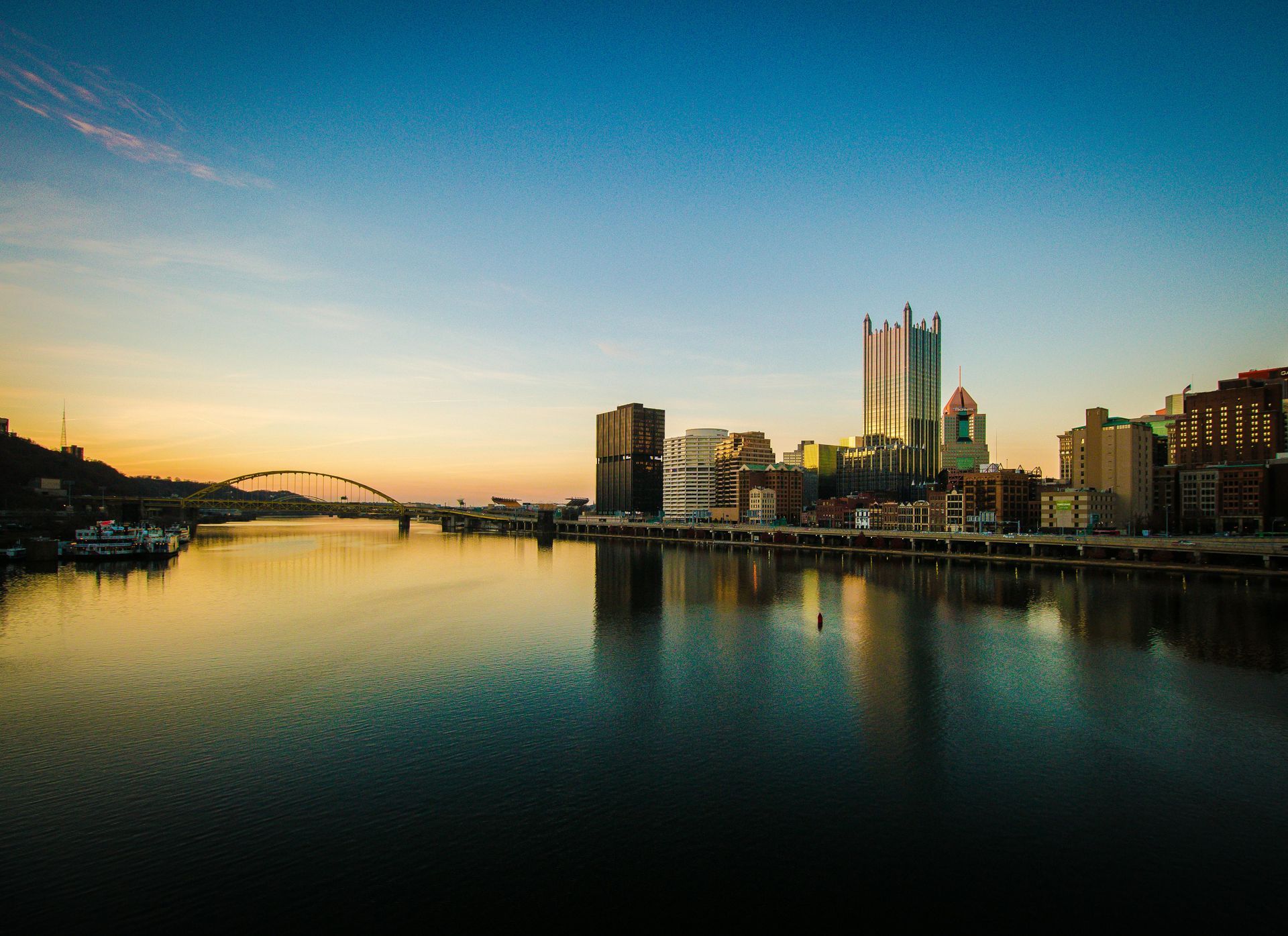 Pittsburgh skyline reflected in a calm river at dawn. Buildings silhouetted against a colorful sky with gold and blue hues.