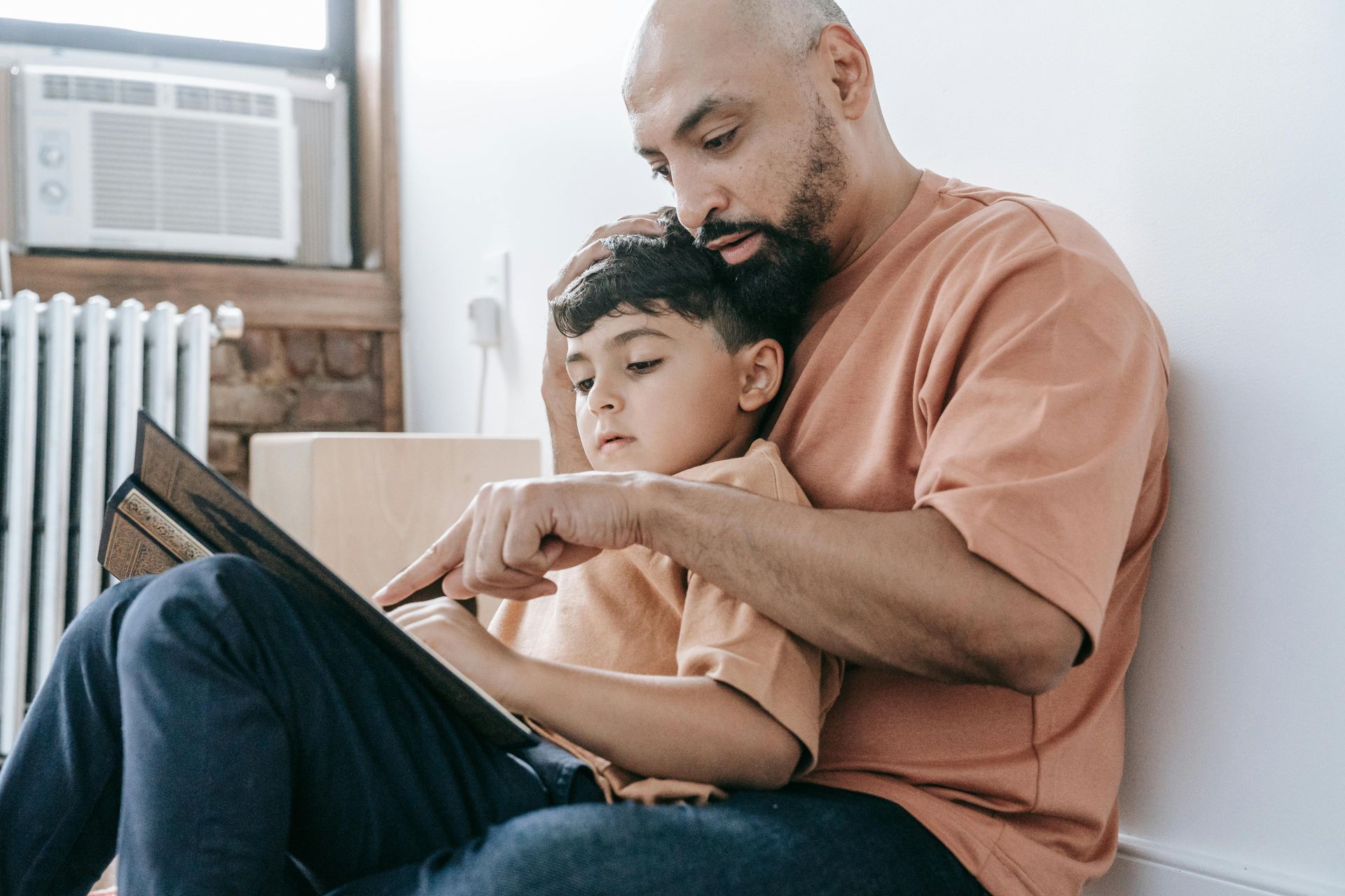 A man is sitting on the floor reading a book to his son.