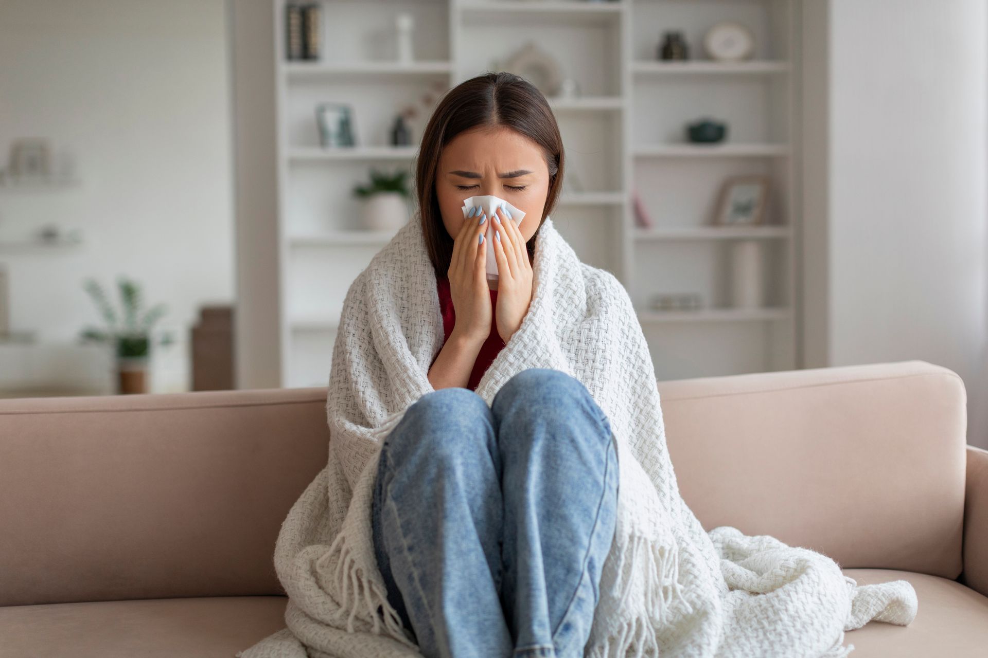 A woman wrapped in a blanket is sitting on a couch blowing her nose.