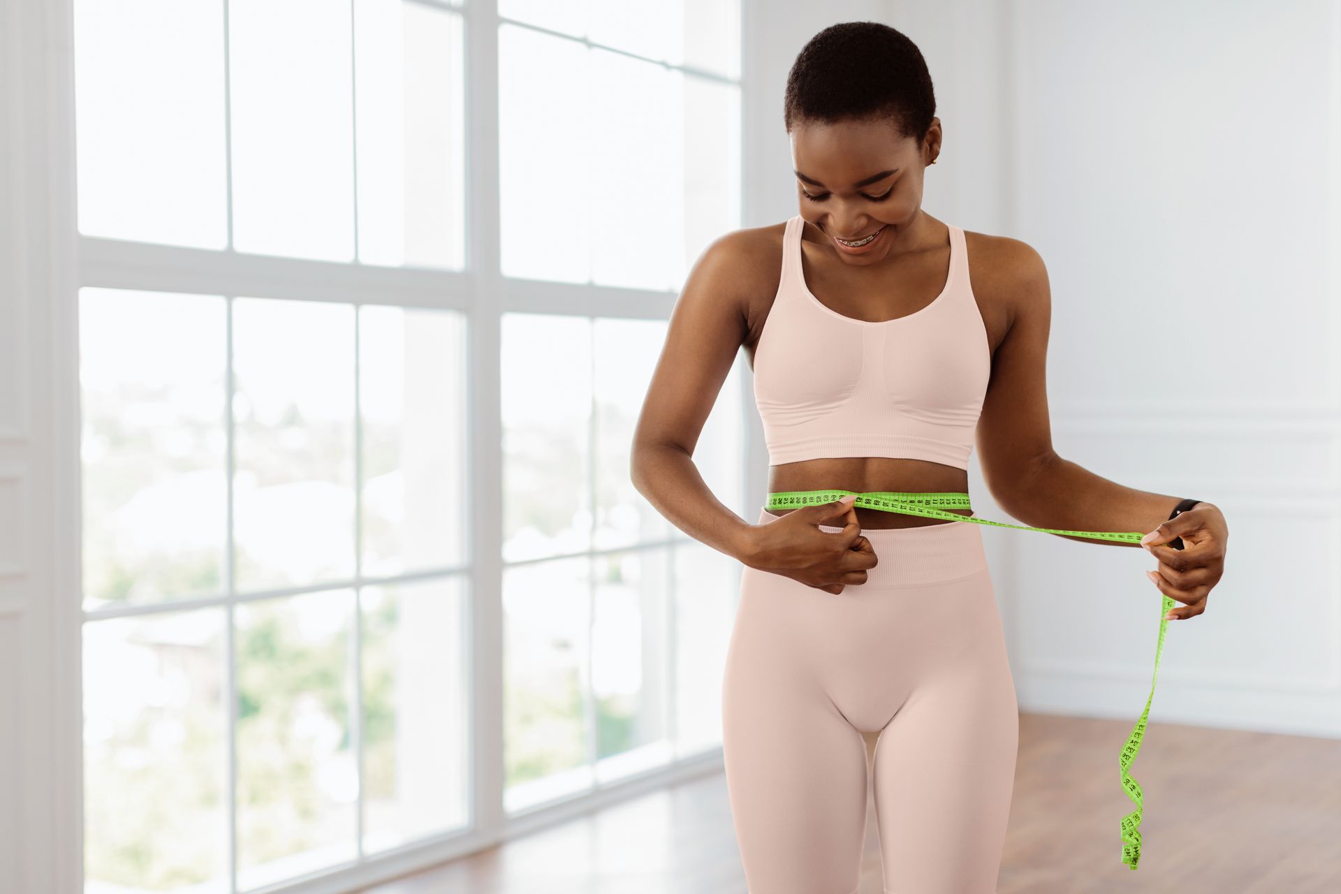 Woman in exercise outfit measuring waist with a tape measure in a bright room.
