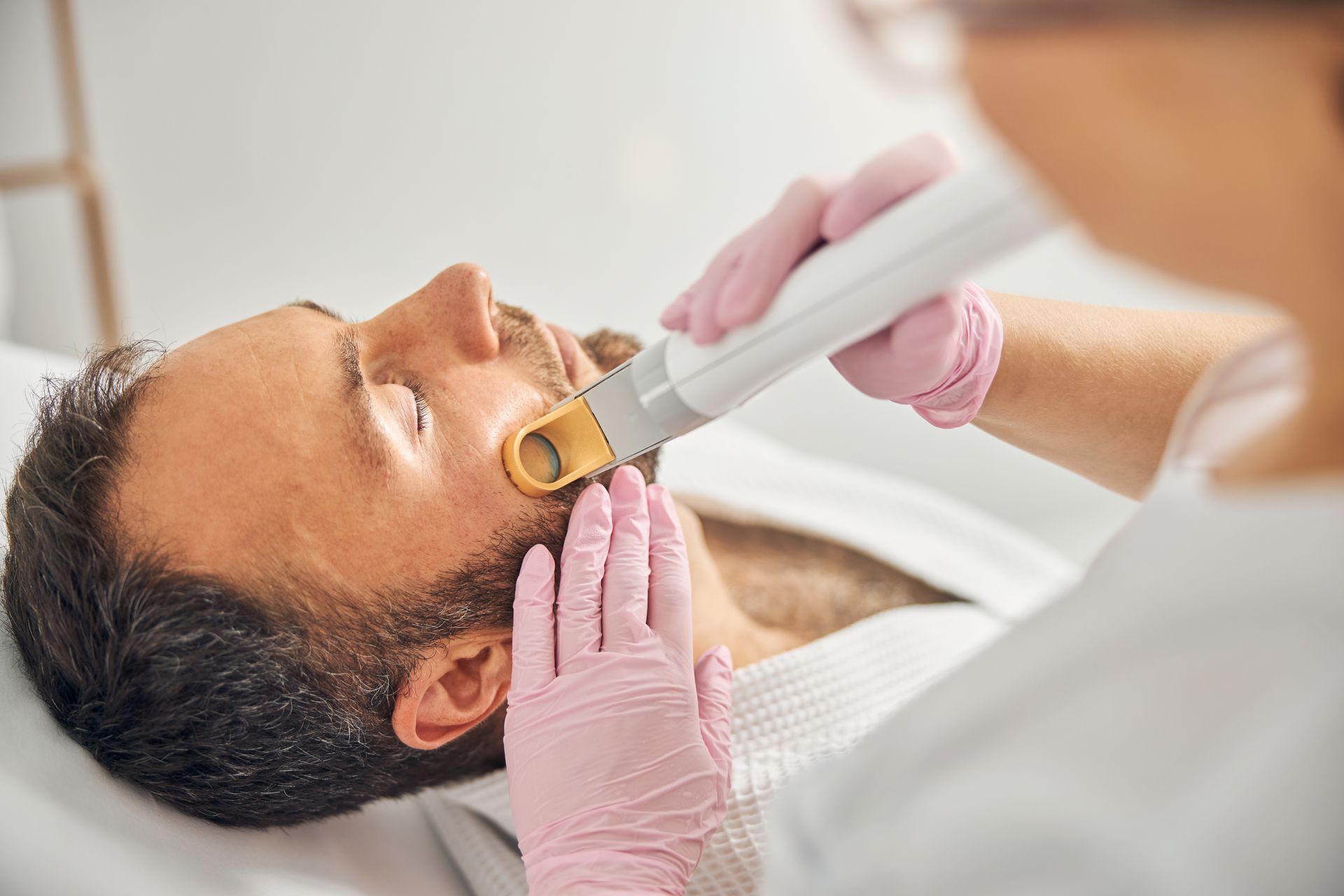 A skincare clinician uses a laser device on a man’s face while wearing pink gloves.