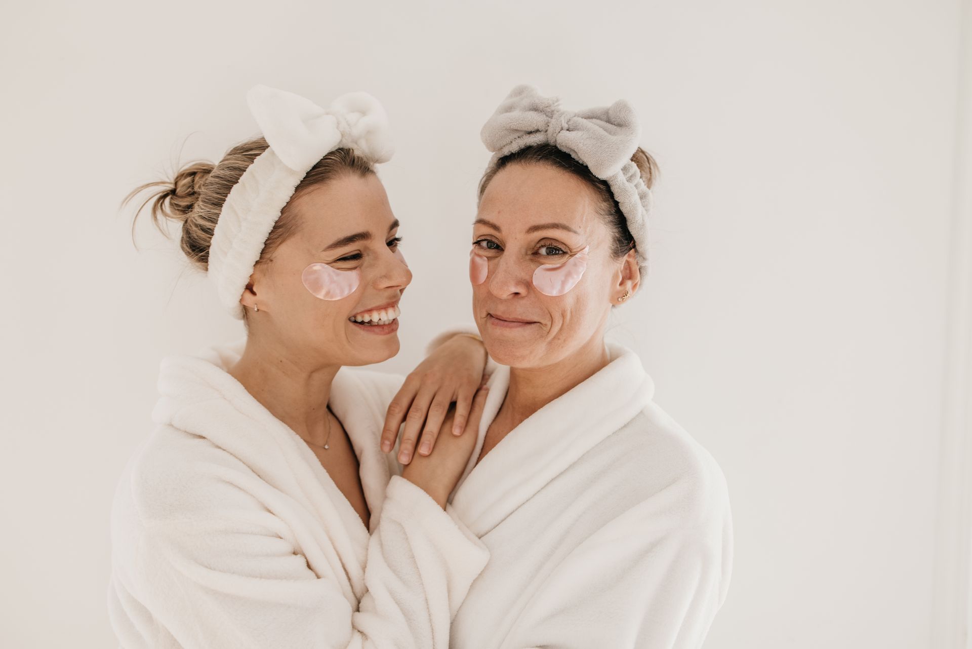 Two women in white bathrobes and headbands smiling together against a light background