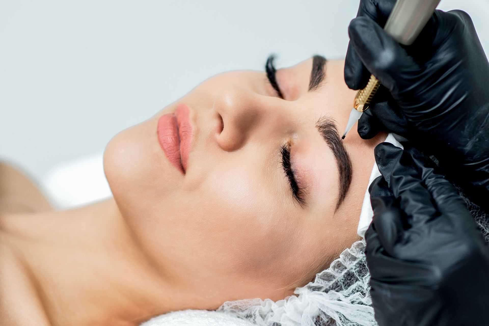 Woman receiving eyebrow microblading with gloved hands and a tattoo machine in a studio setting.